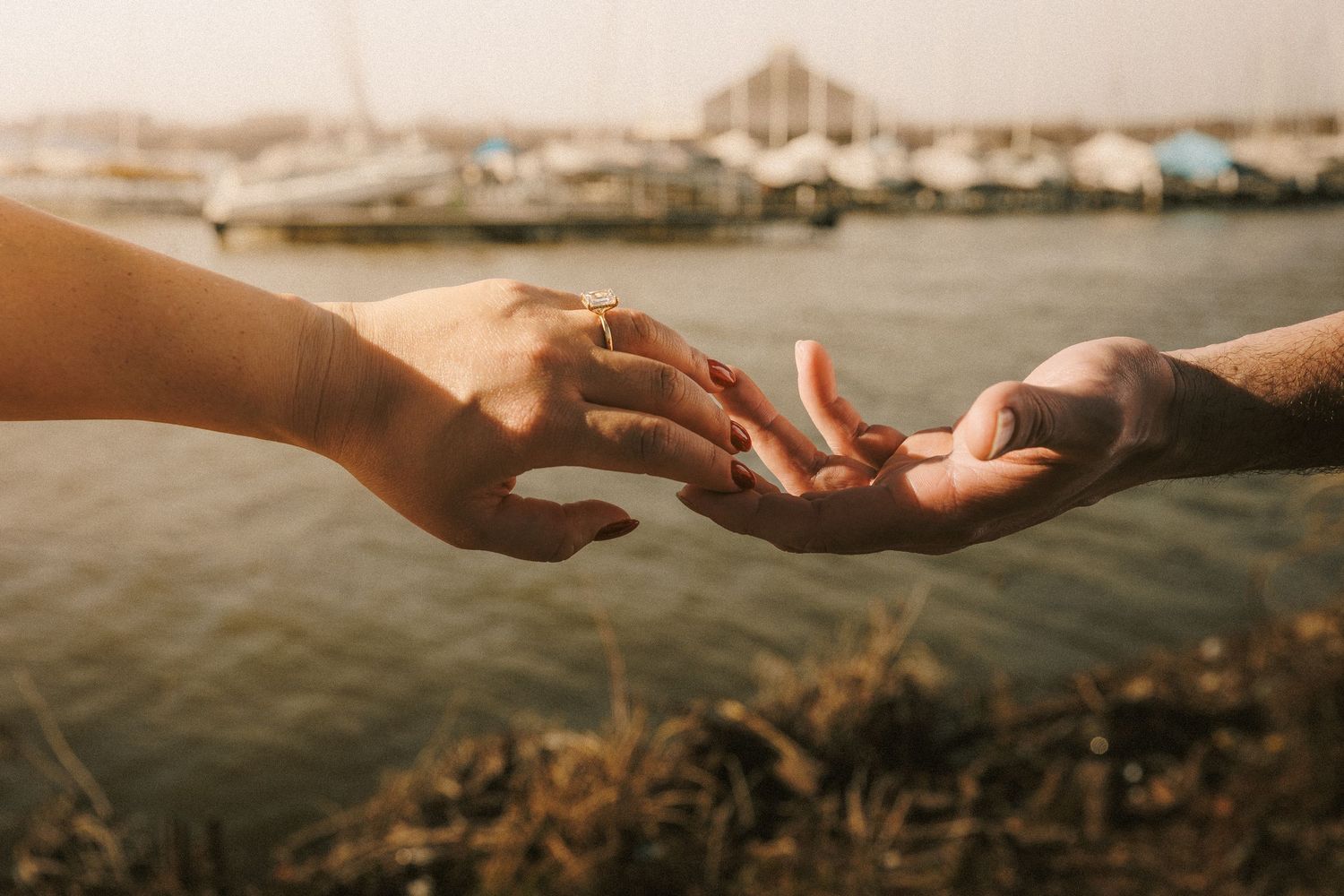 Close-up of hands reaching toward each other against a waterfront backdrop at sunset.