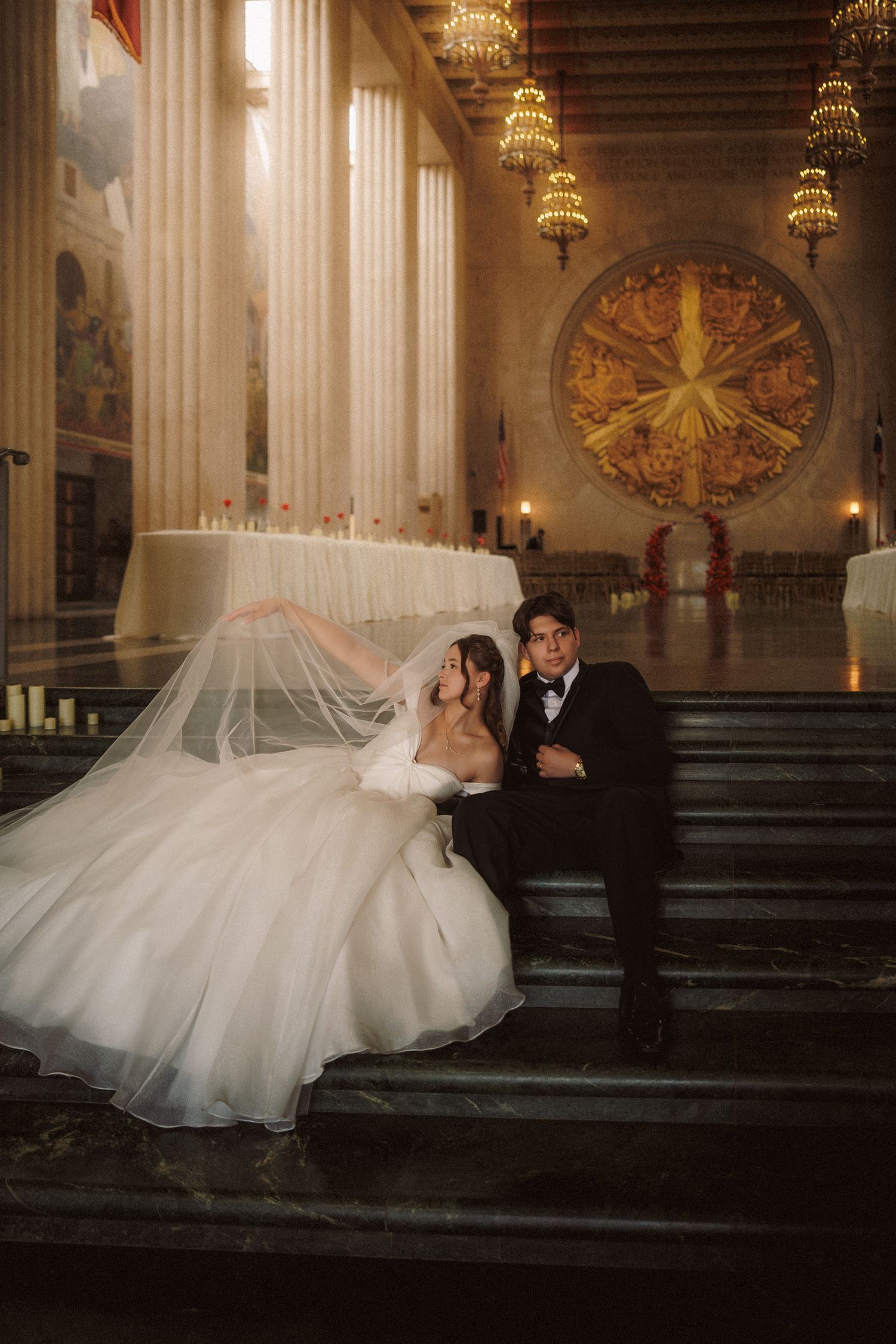 Bride and groom pose elegantly on marble steps inside a grand ballroom with ornate chandeliers and golden decor.