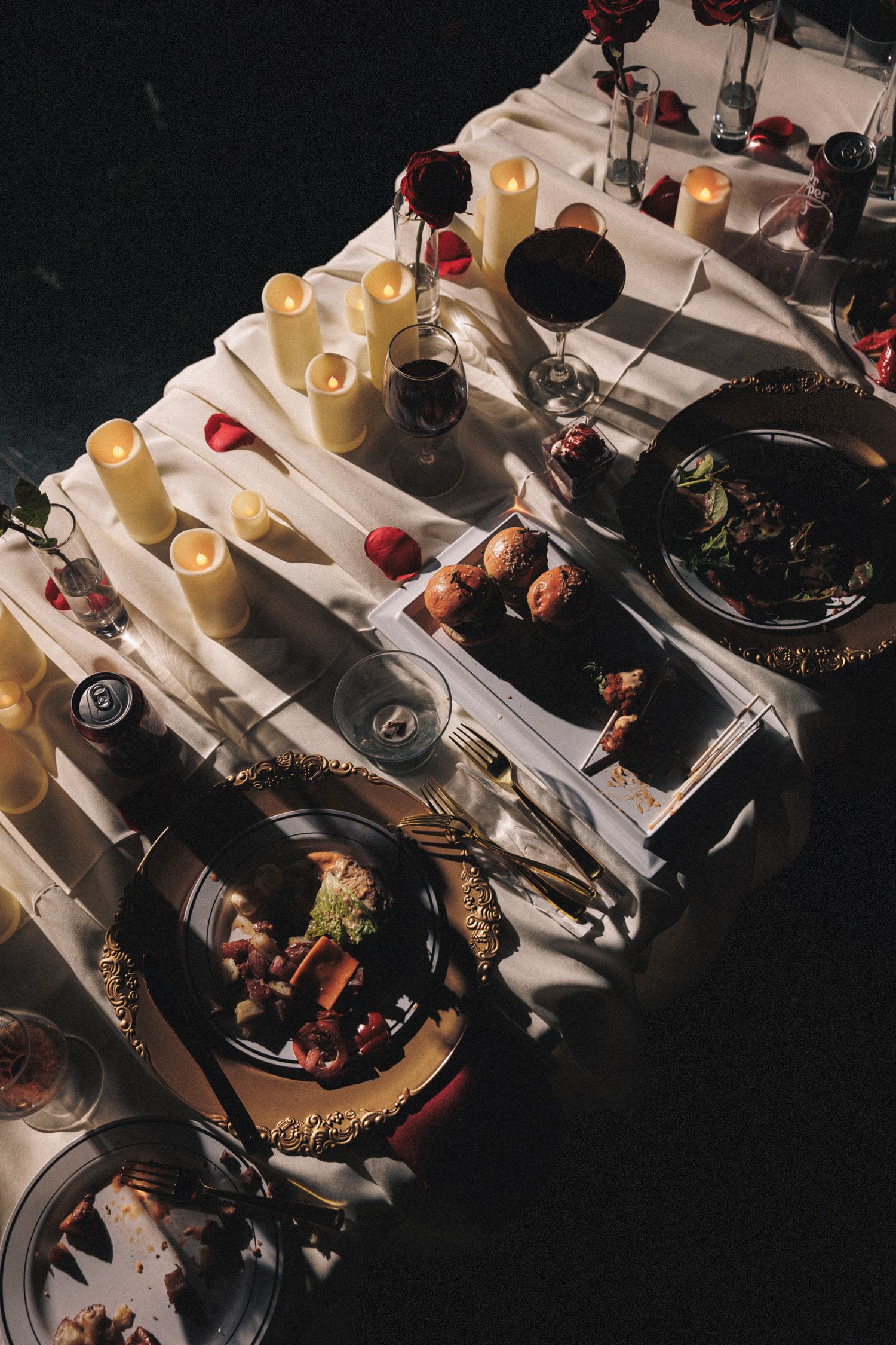 Elegant candlelit dinner table with white linens, pillar candles, red wine, rose petals, and gourmet dishes for a romantic setting.