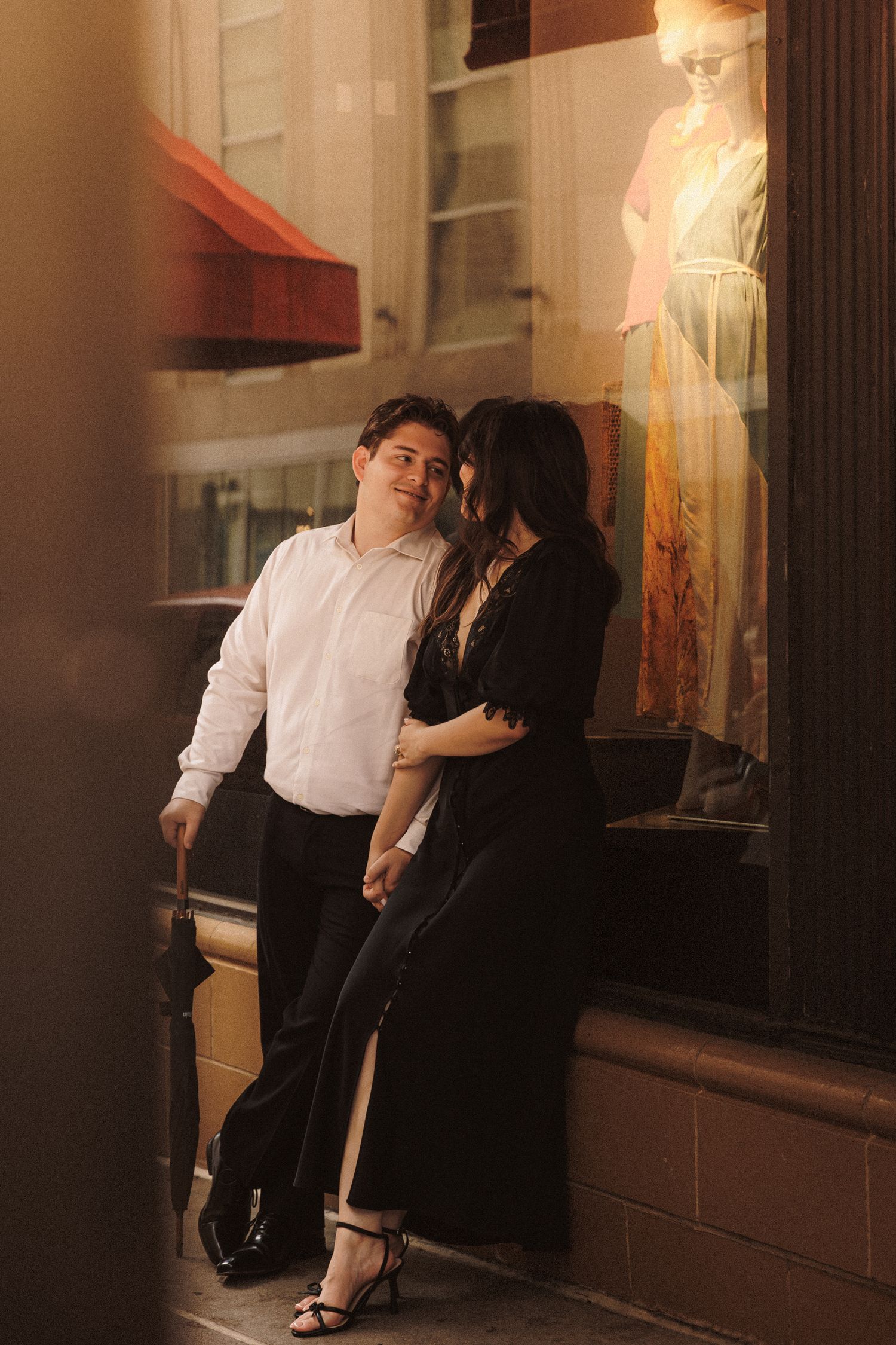 Couple posing romantically outside a boutique storefront at night, man in white shirt and woman in black dress.