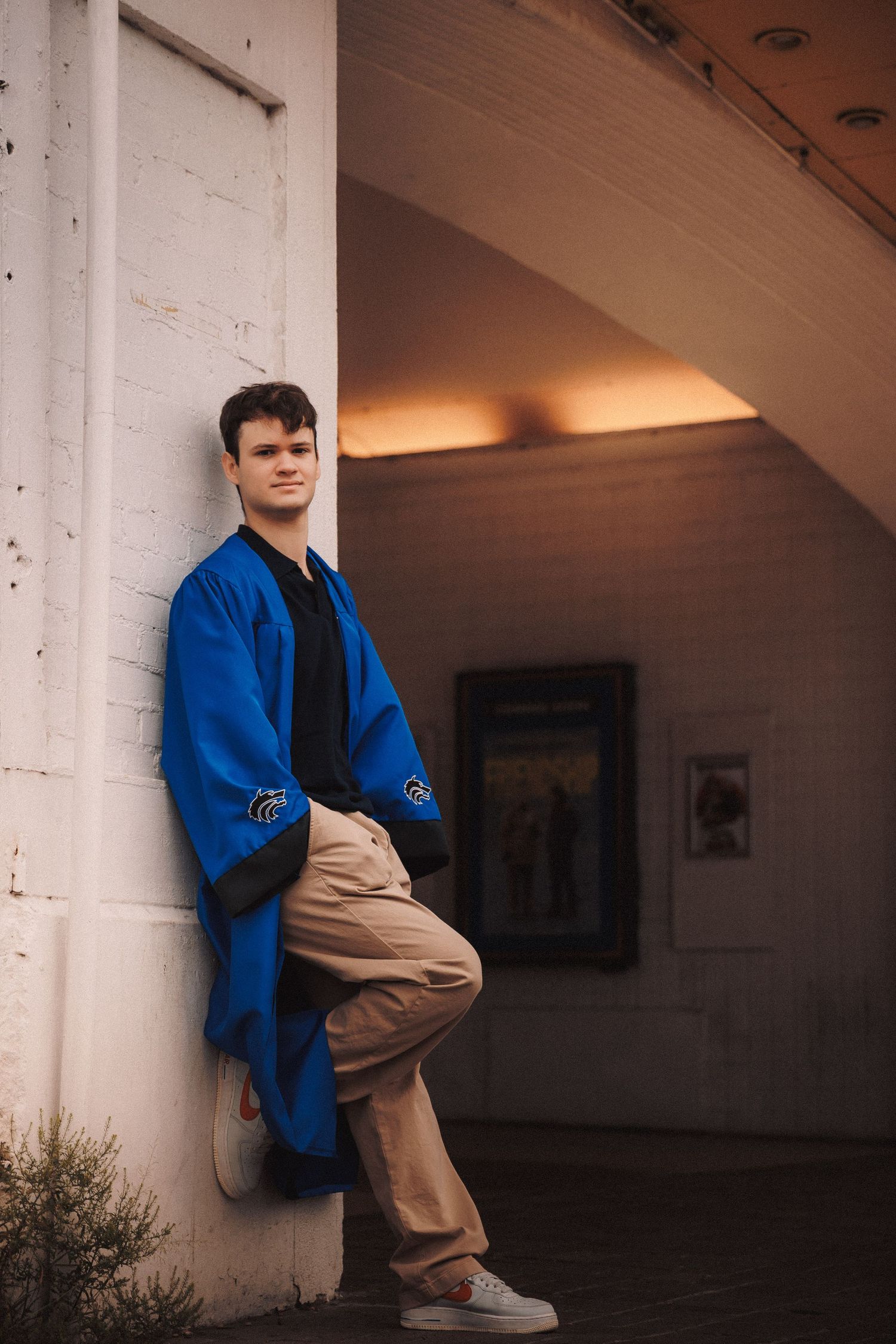 Person in bright blue jacket and khaki pants sits casually against white column in artistic portrait series.