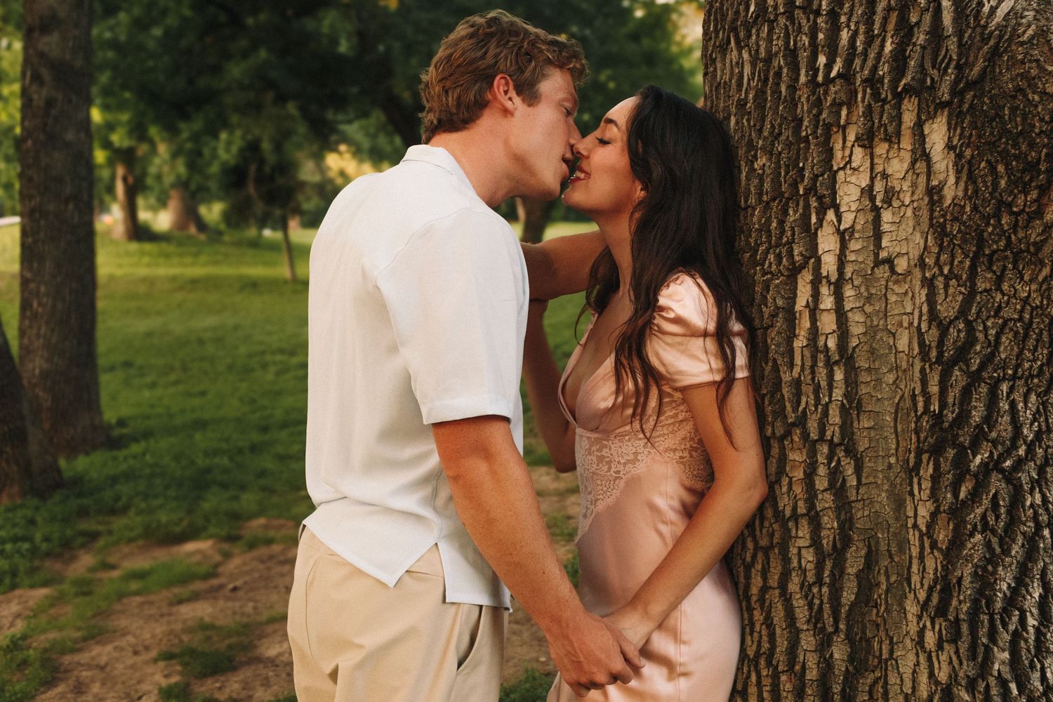 Romantic couple sharing an intimate moment against a tree trunk during sunset in a park.