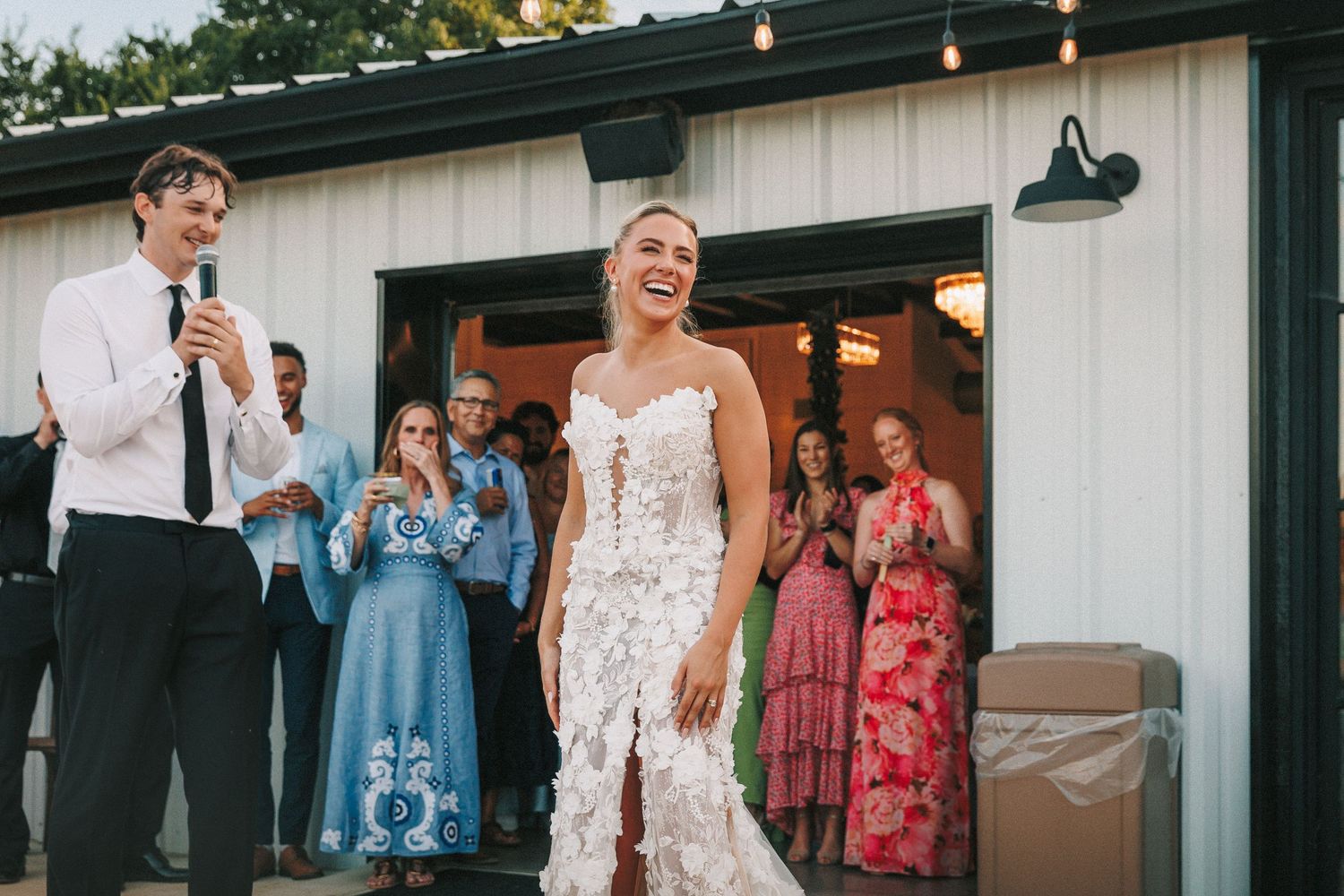 Wedding guests gather outside a white venue building with black trim and string lights at dusk.