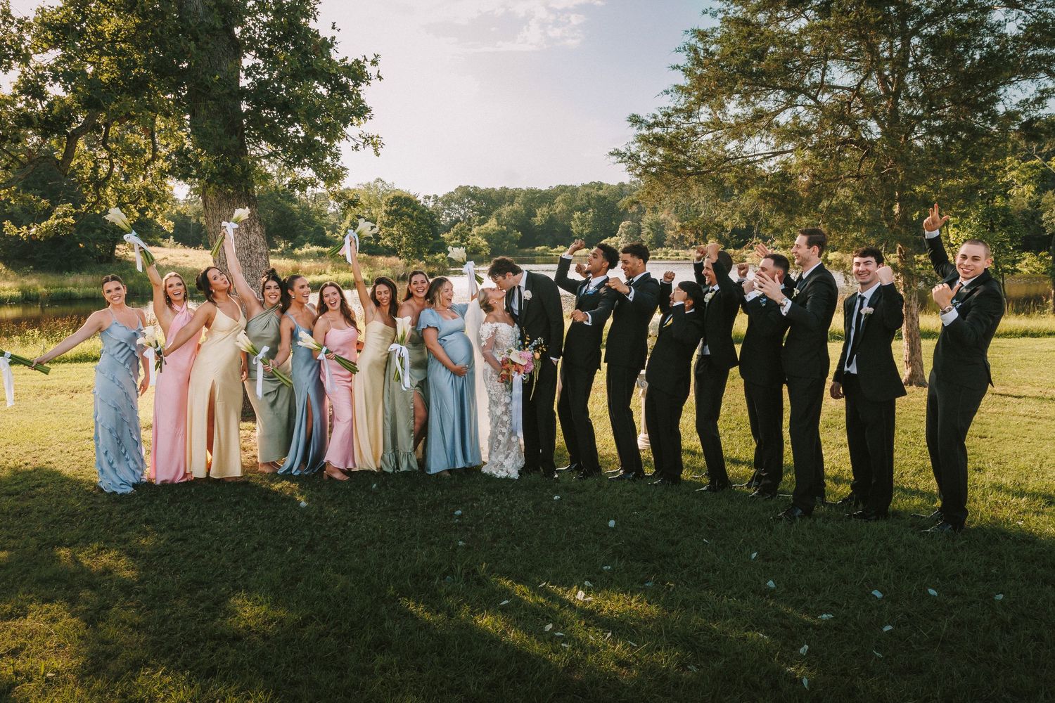 Large wedding party lined up for group photos in a grassy field during golden hour.