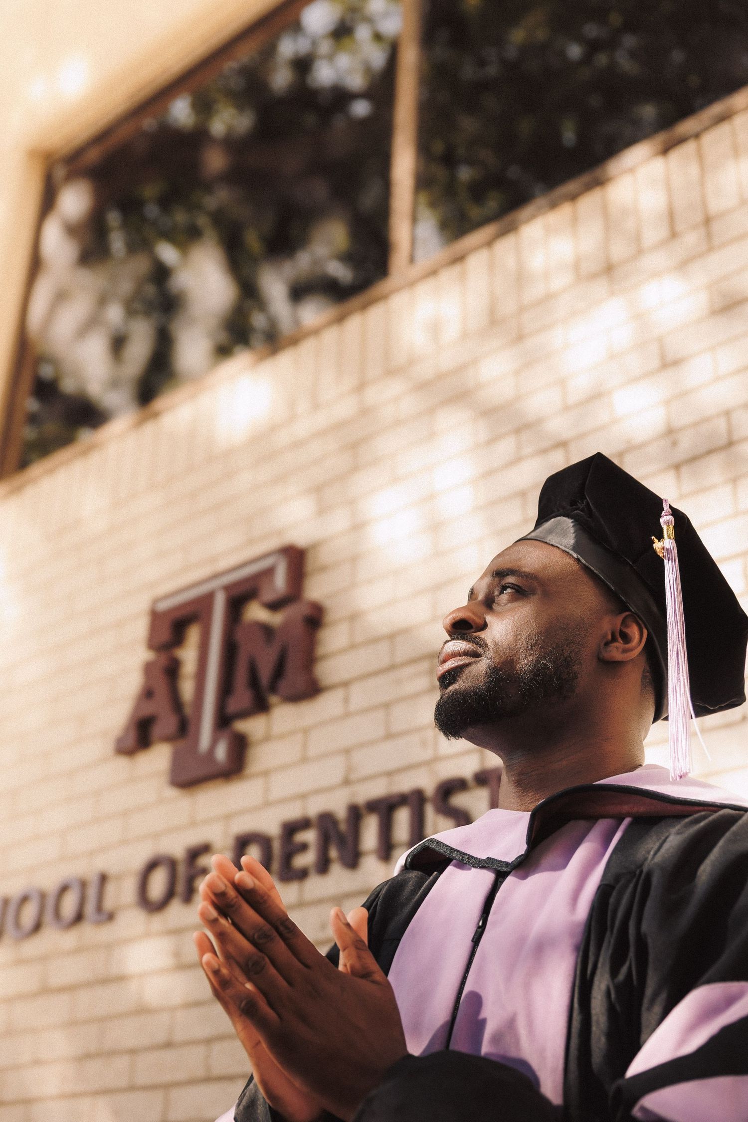 Graduate in black cap and gown with pink stole lifts hands in joyful celebration against campus wall background.