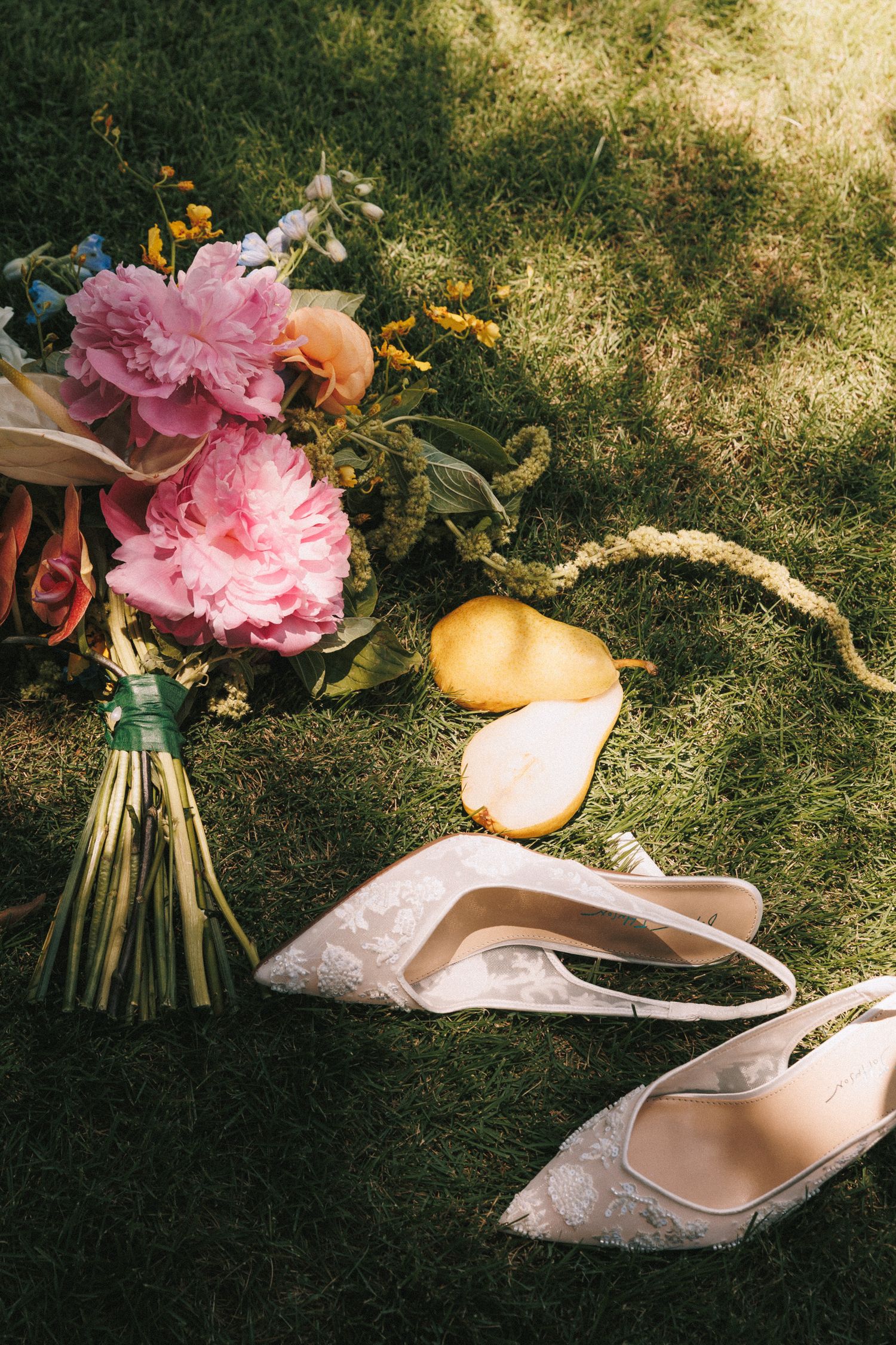 Pink peony flowers and elegant beige bridal shoes arranged on grass in natural sunlight.