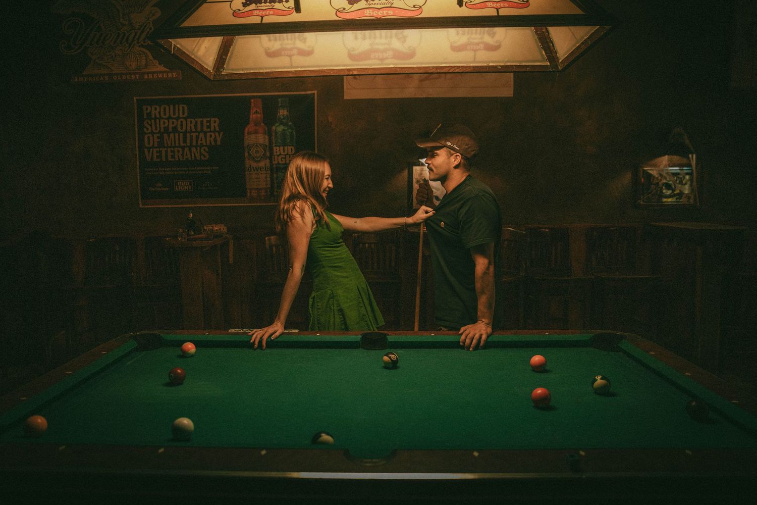 People playing pool in a dimly lit billiards room with multiple tables and overhead lighting.