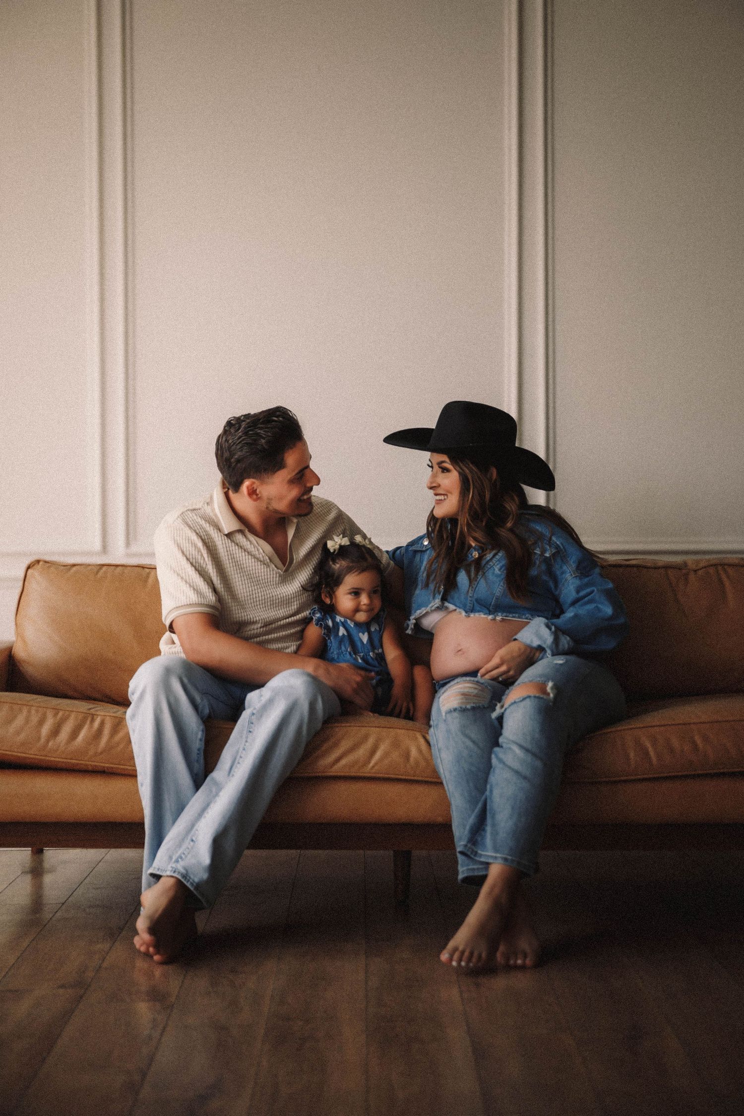 A couple sits barefoot on a brown leather couch while smiling and sharing a tender family moment together.