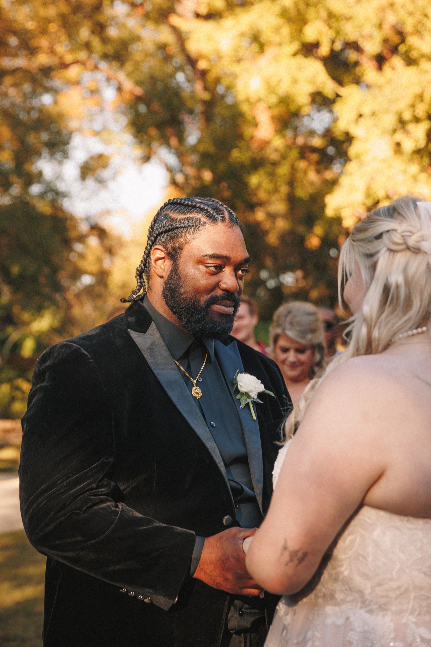 Groom in black velvet suit holds bride's hands during outdoor fall wedding ceremony.