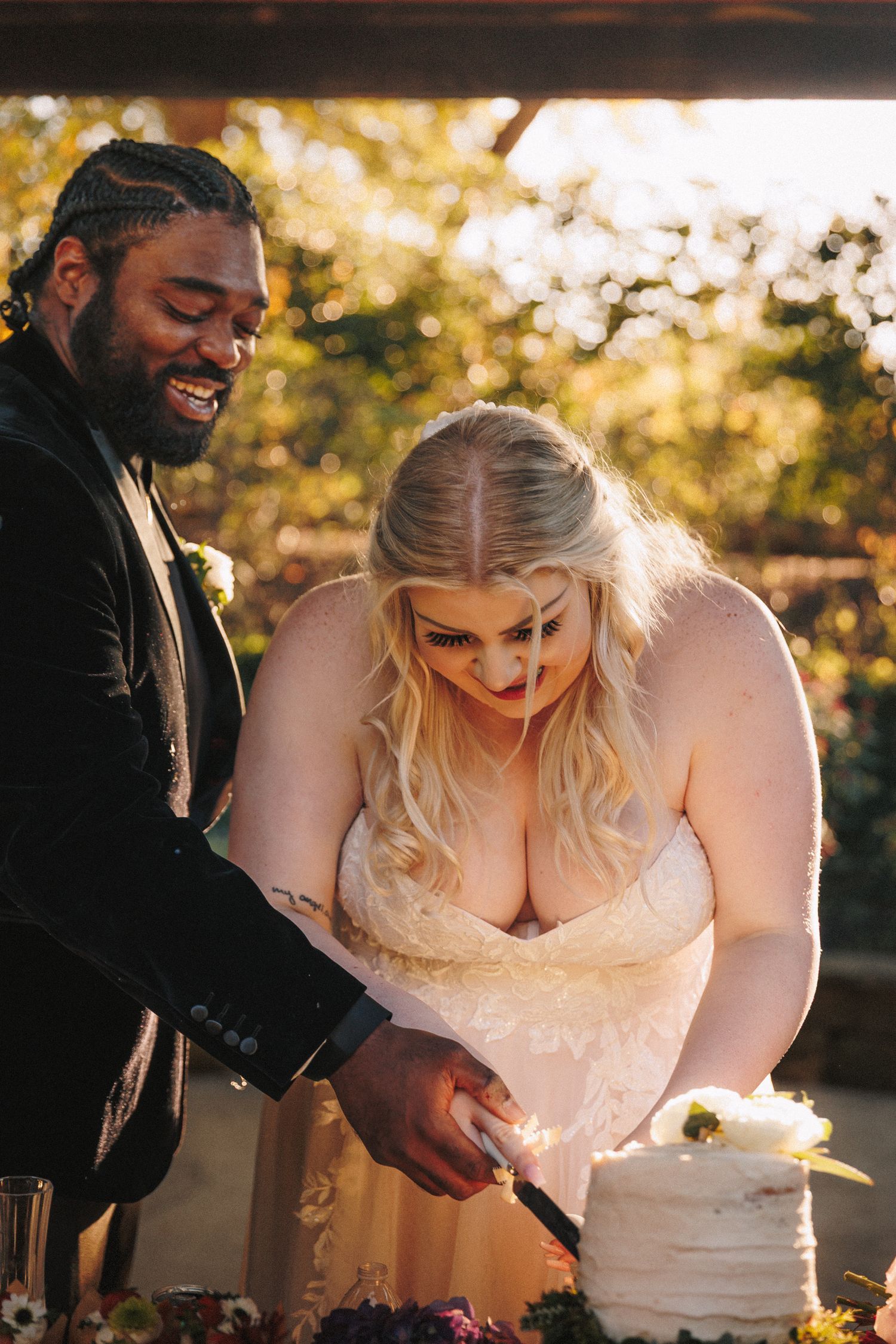 Interracial newlywed couple smiles while cutting white wedding cake at outdoor reception.