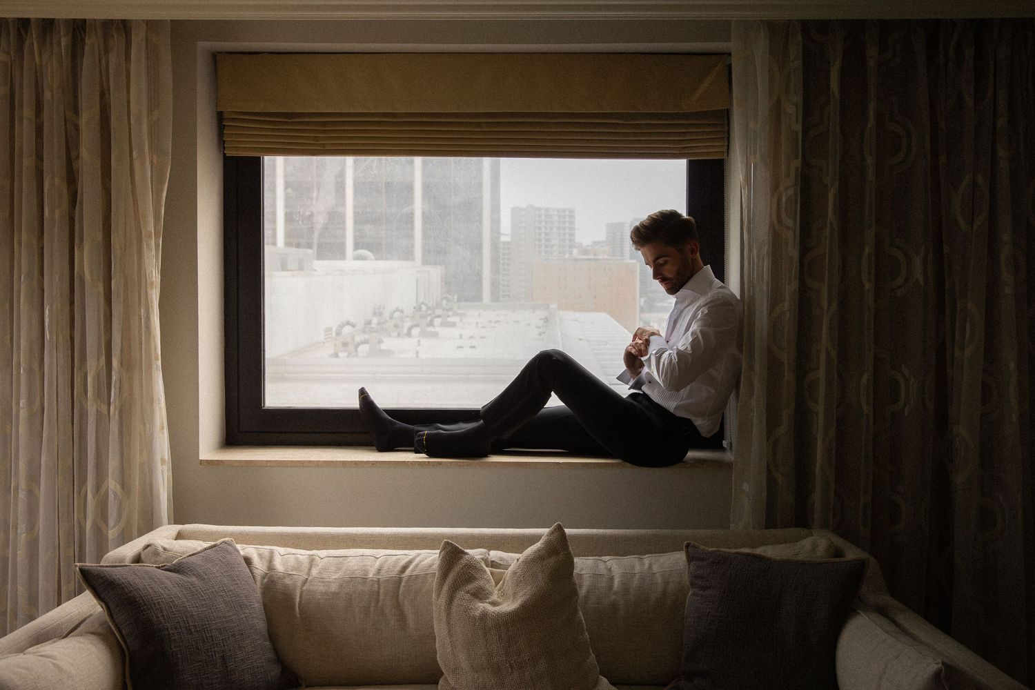 Groom sitting on hotel window sill in white dress shirt and black pants, preparing for wedding day.