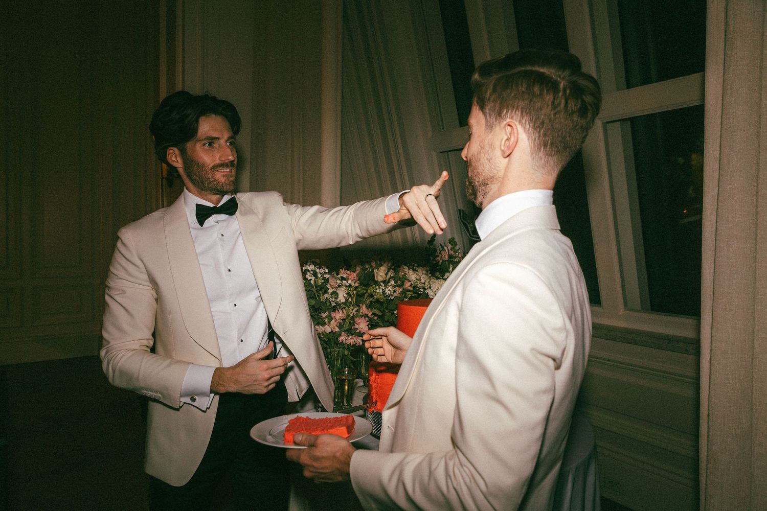 Groom in ivory tuxedo playfully feeds wedding cake to partner at elegant reception.