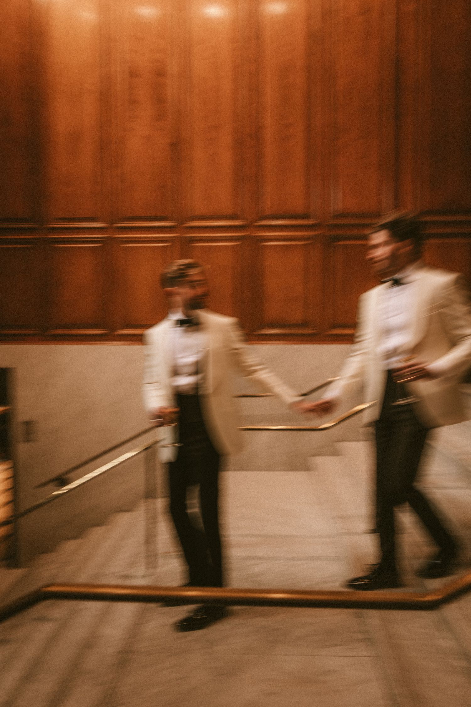 Motion blur artistic photo of two grooms in white tuxedos holding hands on grand staircase.