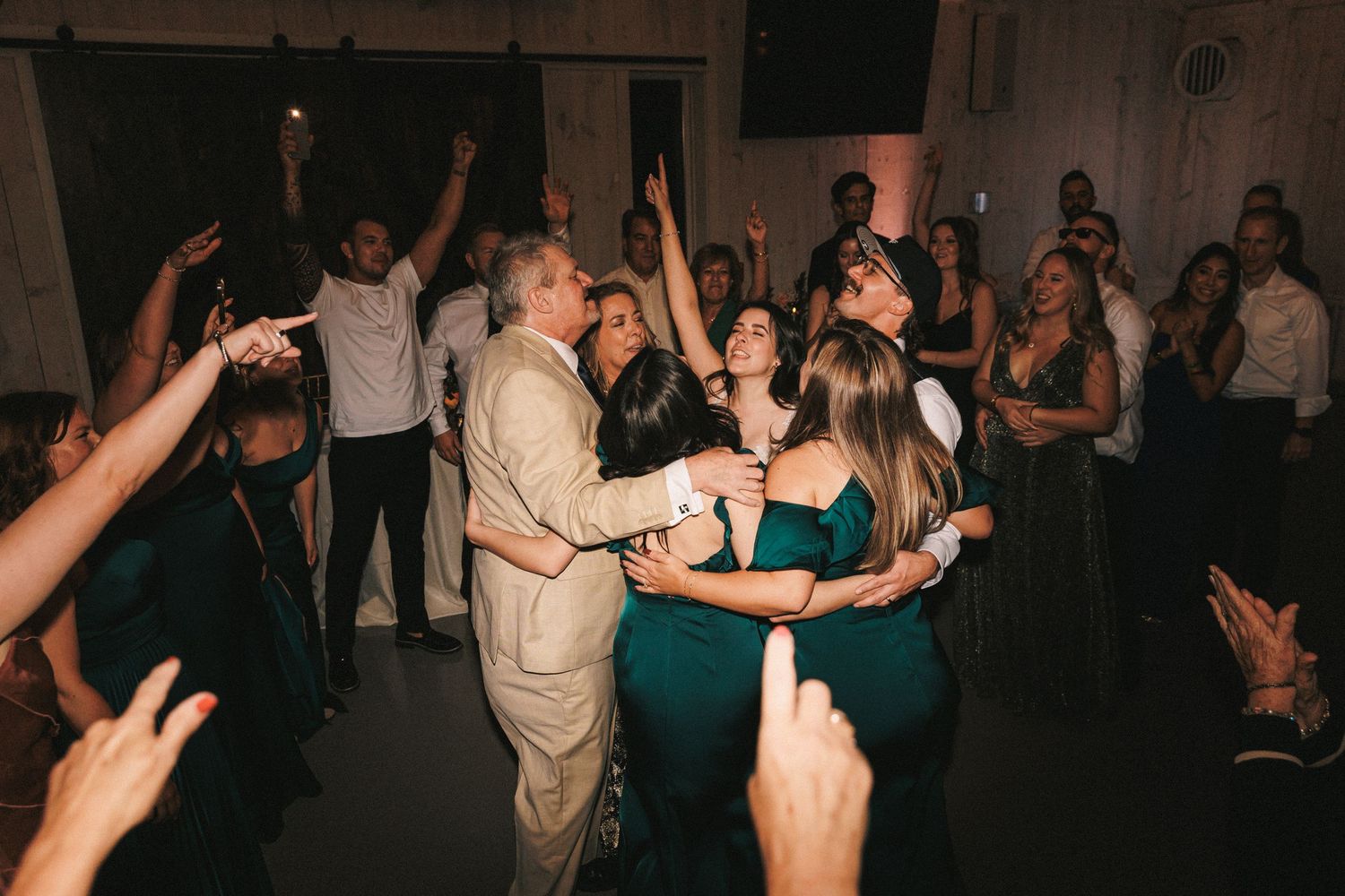 A joyful group of wedding guests in formal attire dance together on a crowded reception dance floor.