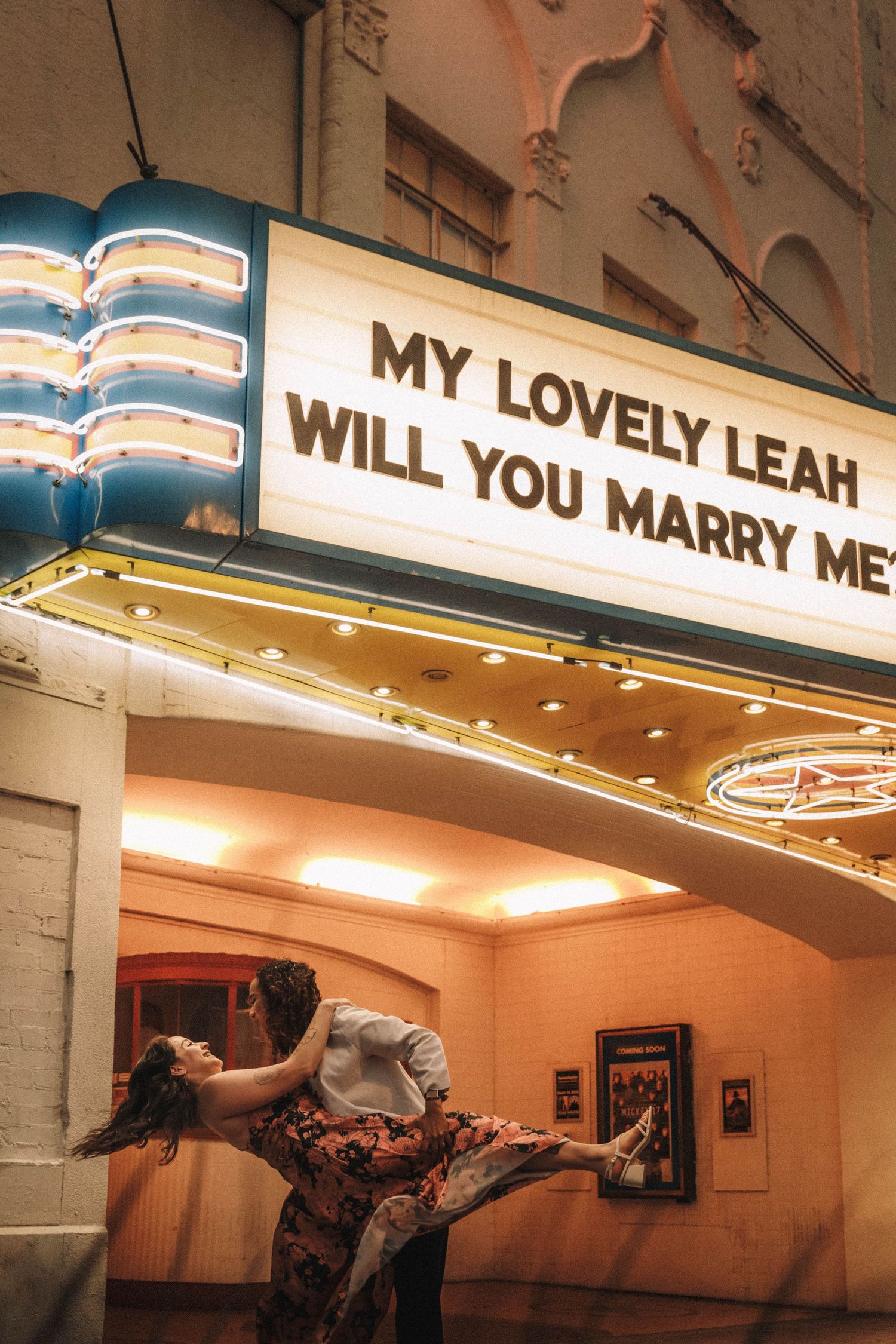 Couple shares romantic dip under glowing theater marquee reading My Lovely Leah Will You Marry Me at night.