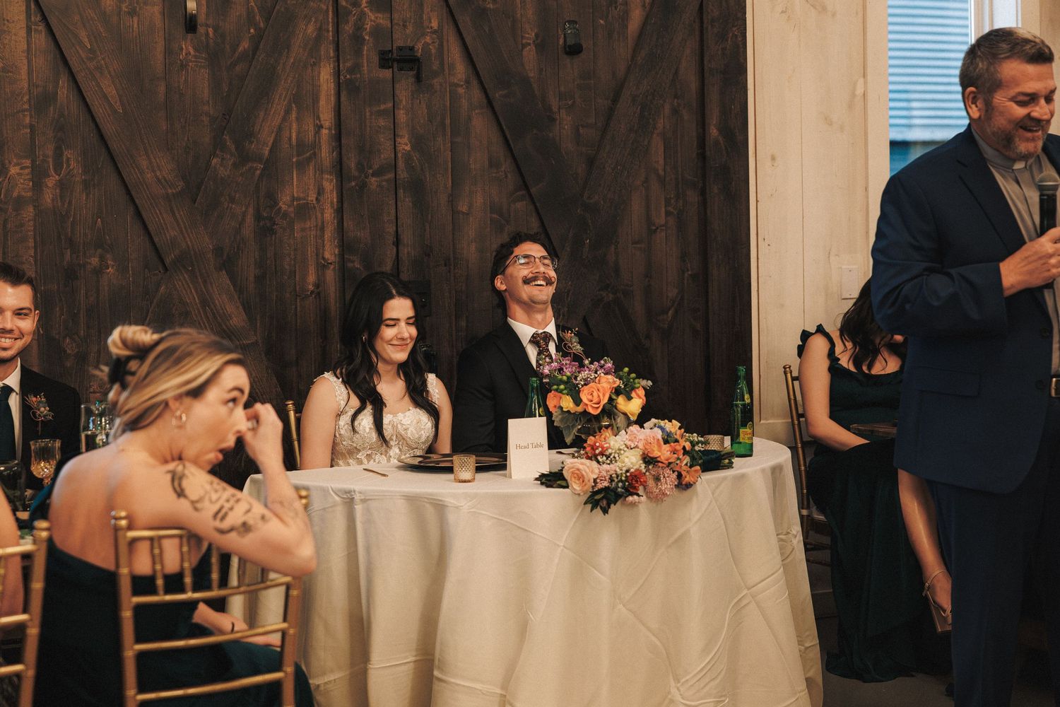 Bride and groom laugh joyfully at sweetheart table during wedding reception speech in rustic barn venue.