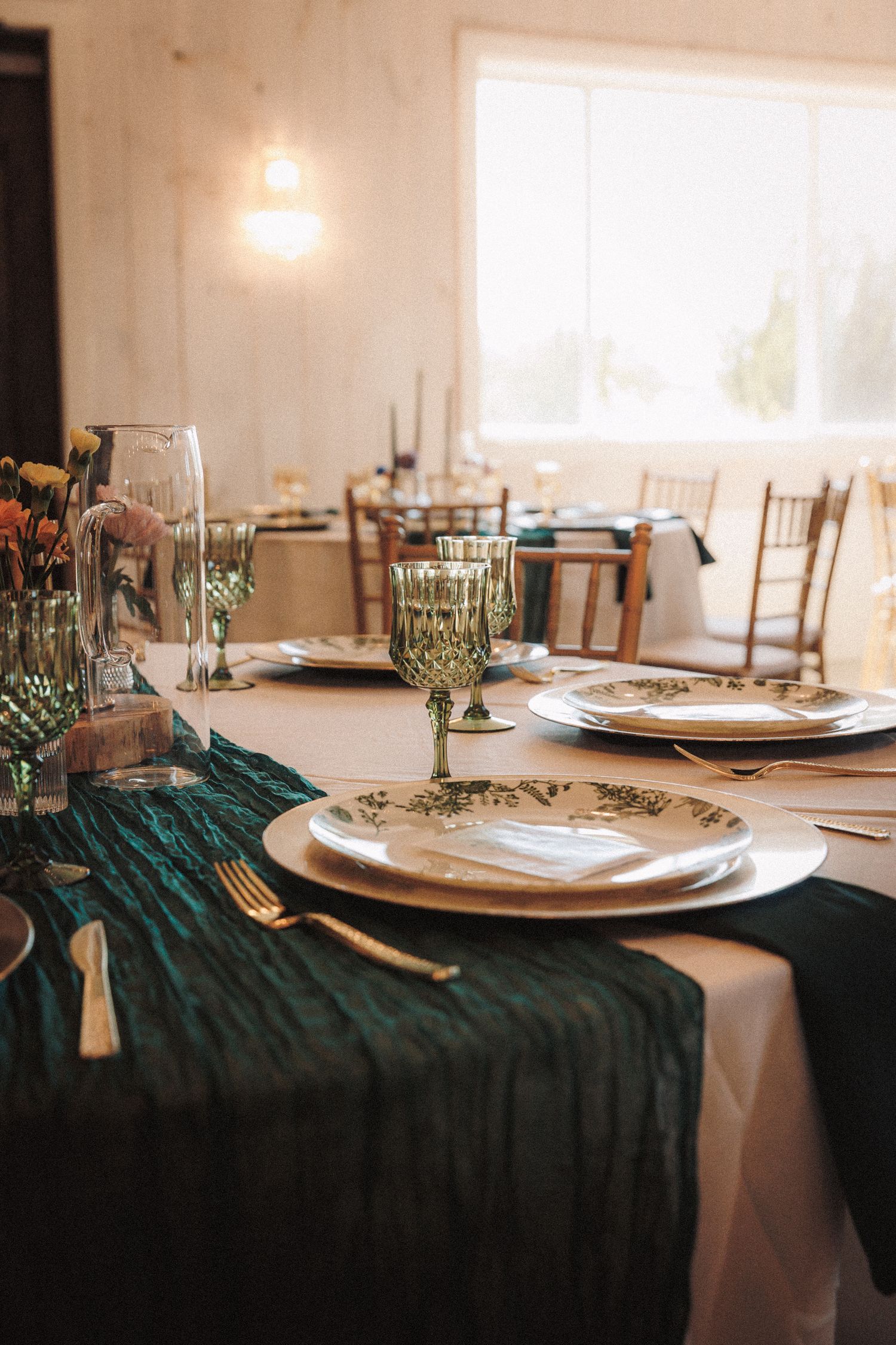 Wedding reception table setting with teal velvet runner, green goblets, gold flatware, and white floral plates.