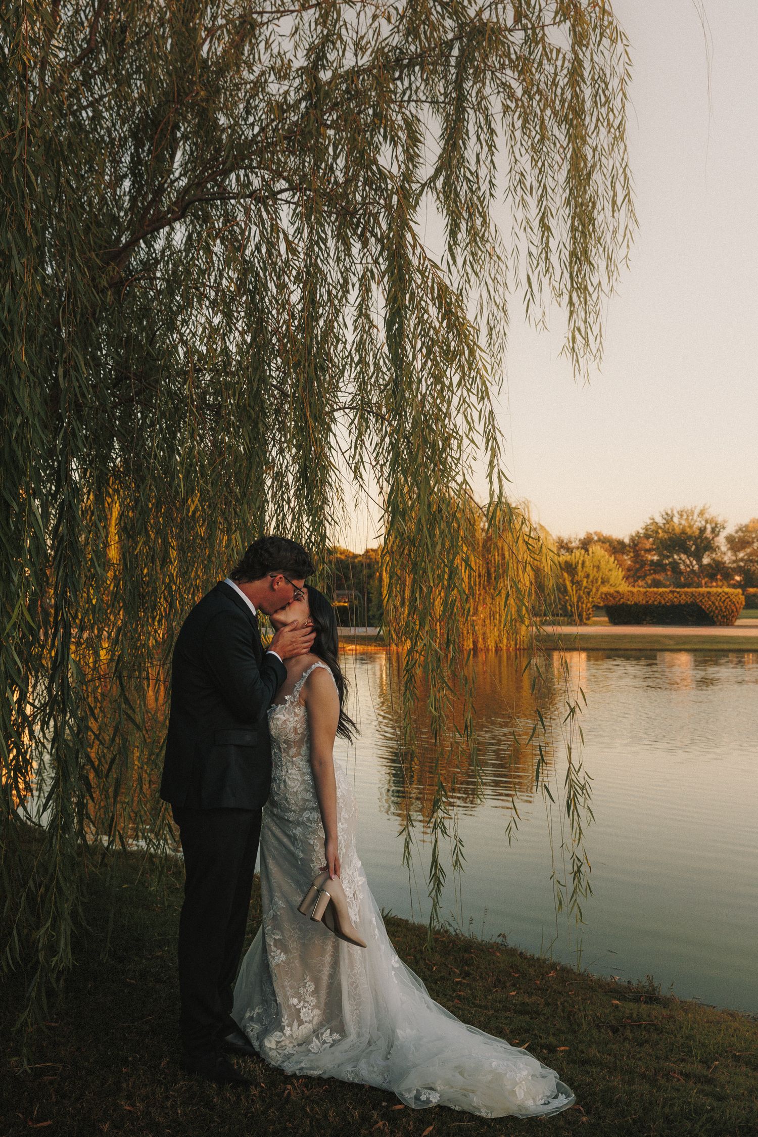 Bride and groom embrace under weeping willow tree beside lake at golden sunset after wedding.