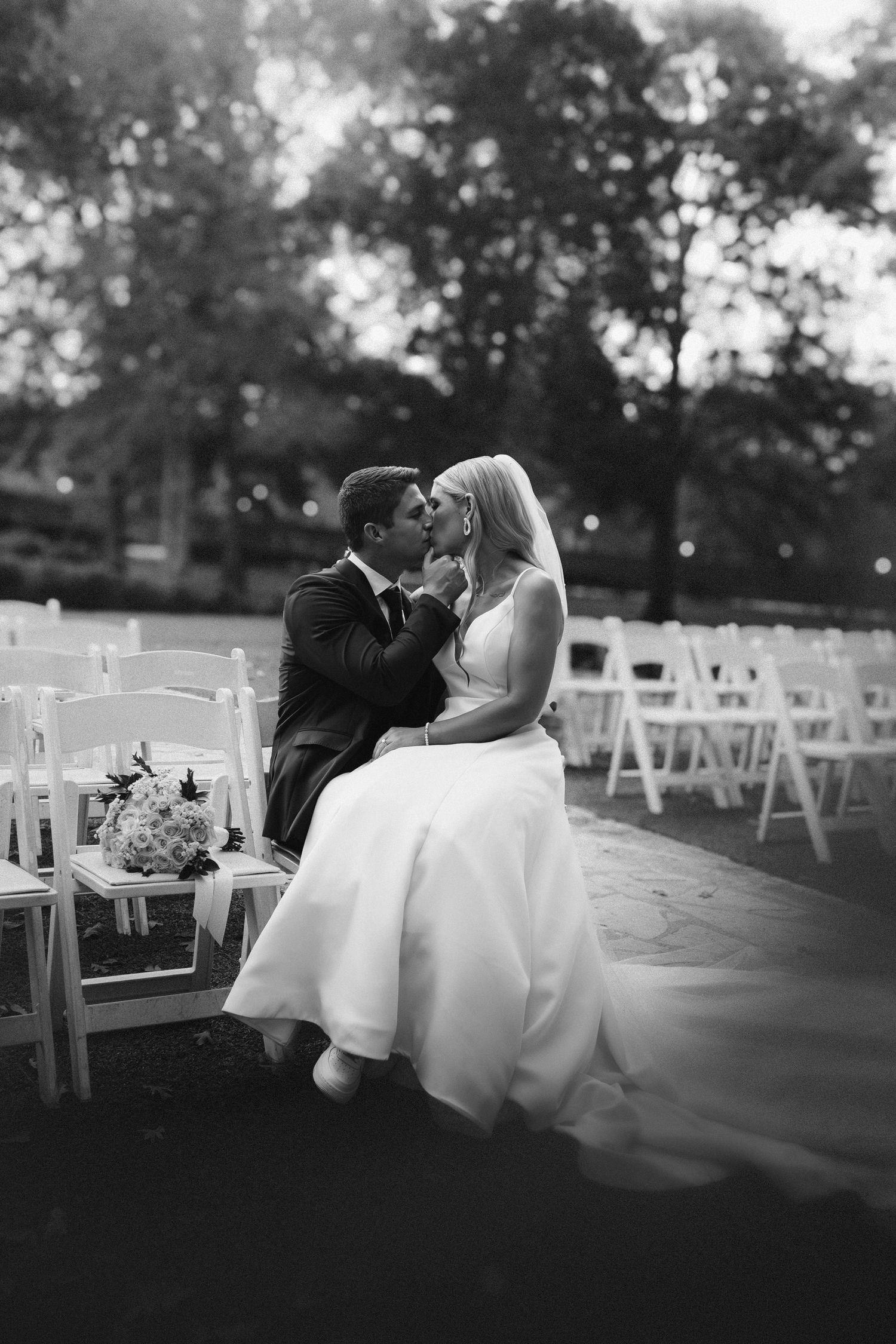 Black and white photo of a bride and groom sharing a kiss while seated among white chairs at an outdoor wedding ceremony.
