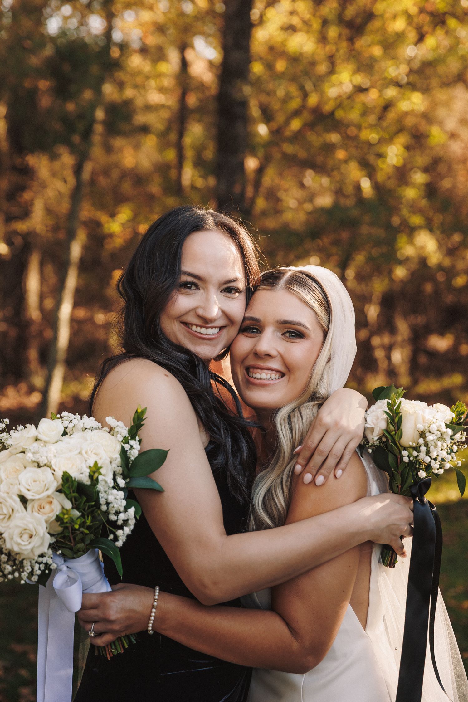 Bride in white gown and bridesmaid in black dress hugging and smiling with white rose bouquets in autumn setting.