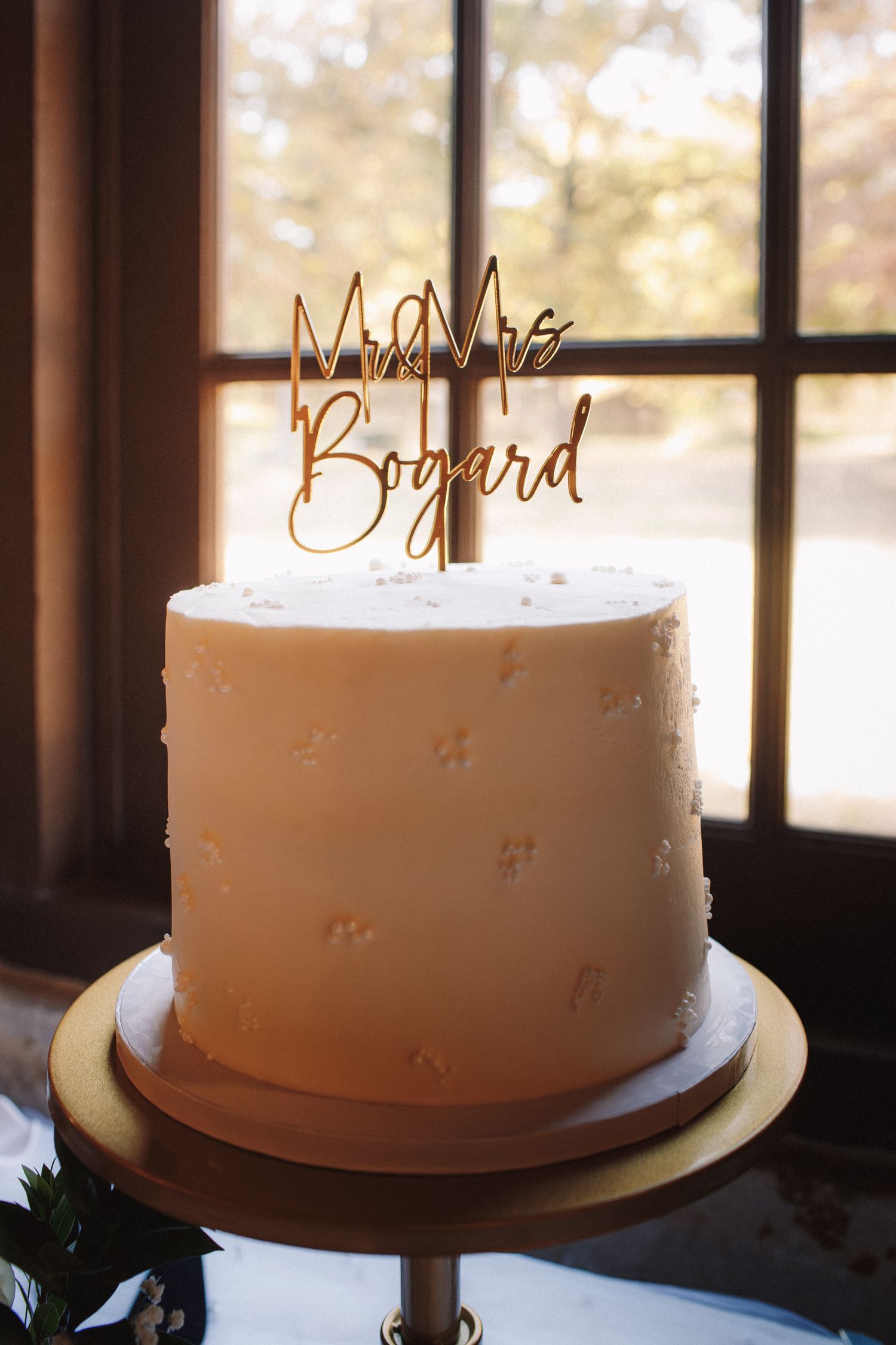 Elegant single-tier white wedding cake with floral texture and gold Mr & Mrs Bogard cake topper on wooden stand.