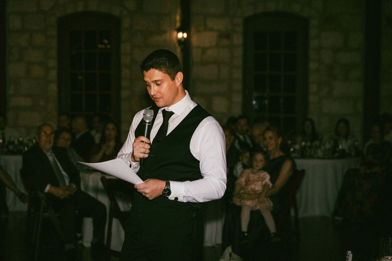 A groomsman in a black vest delivers a heartfelt speech at an elegant wedding reception.