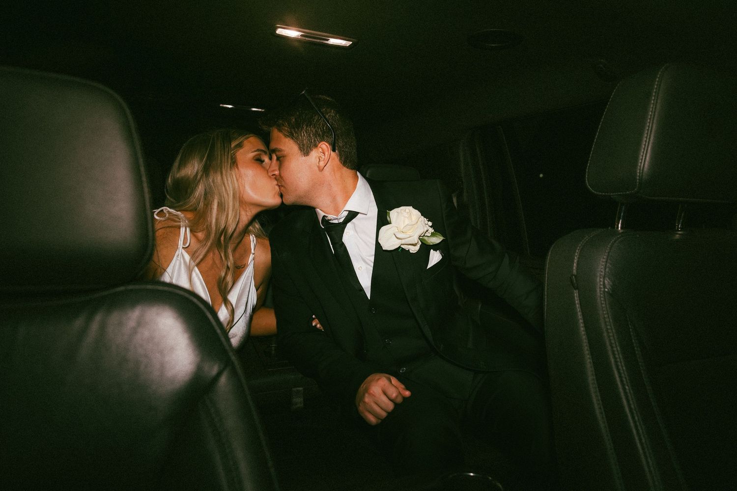 A bride and groom share a kiss in the back of a dark limousine following their wedding ceremony.