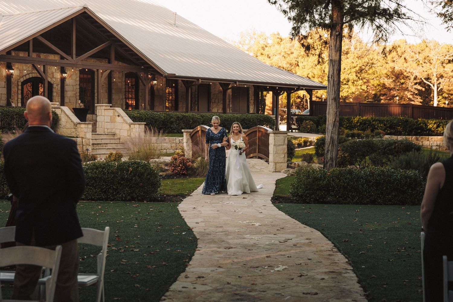 Bride in white gown walks down stone path aisle escorted by woman in blue dress.