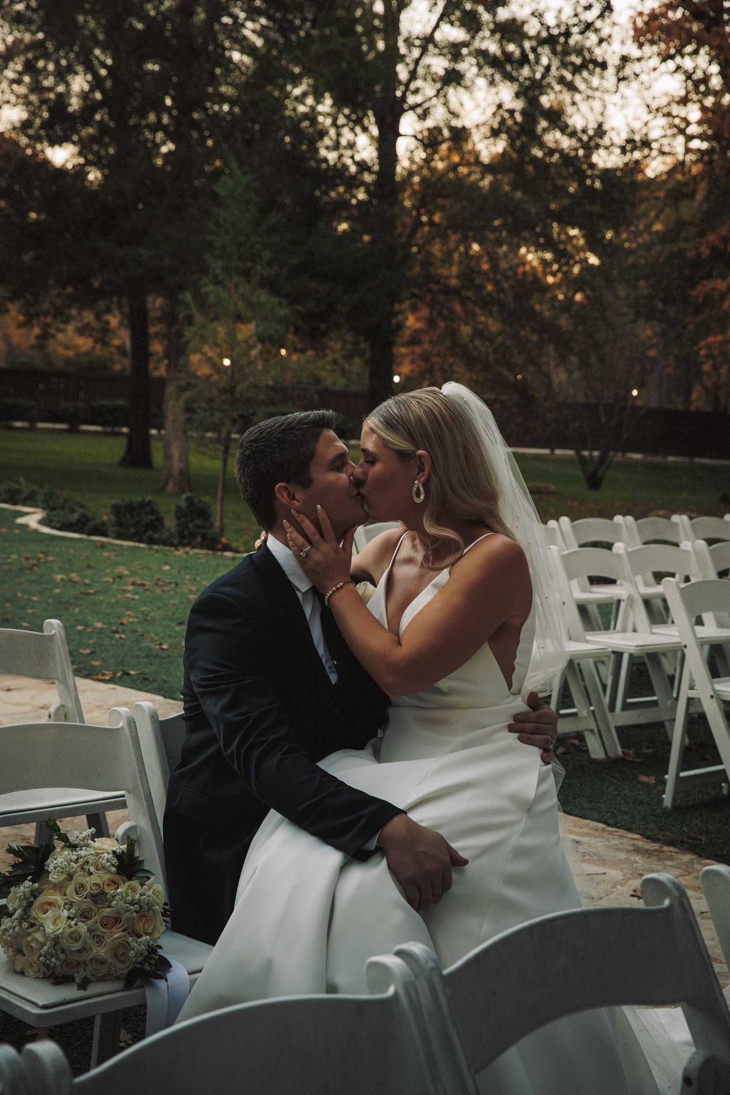 A bride and groom share a romantic kiss among white chairs at their outdoor autumn wedding ceremony at dusk.