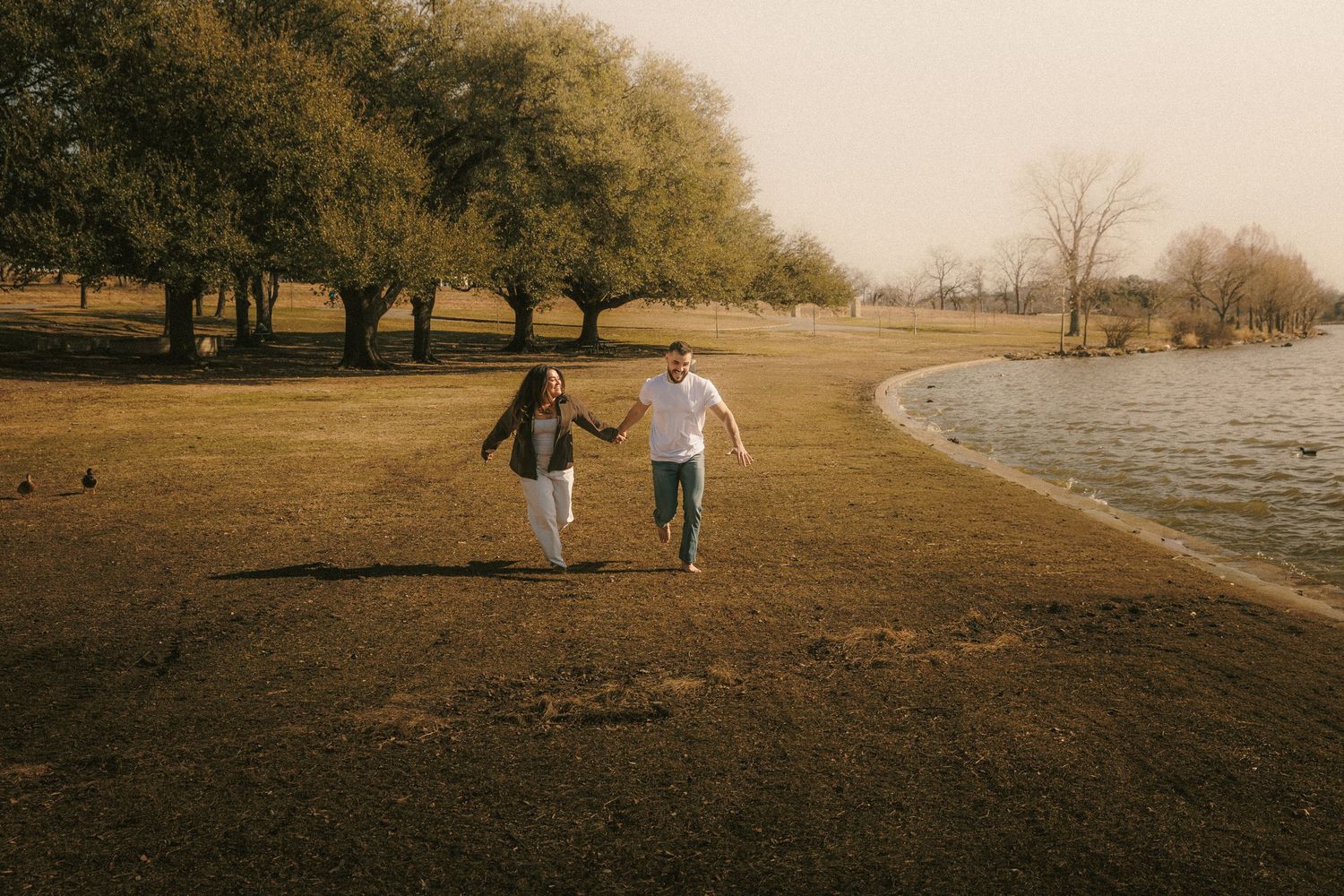 A couple running hand in hand along a lakeside path with autumn trees in the background.