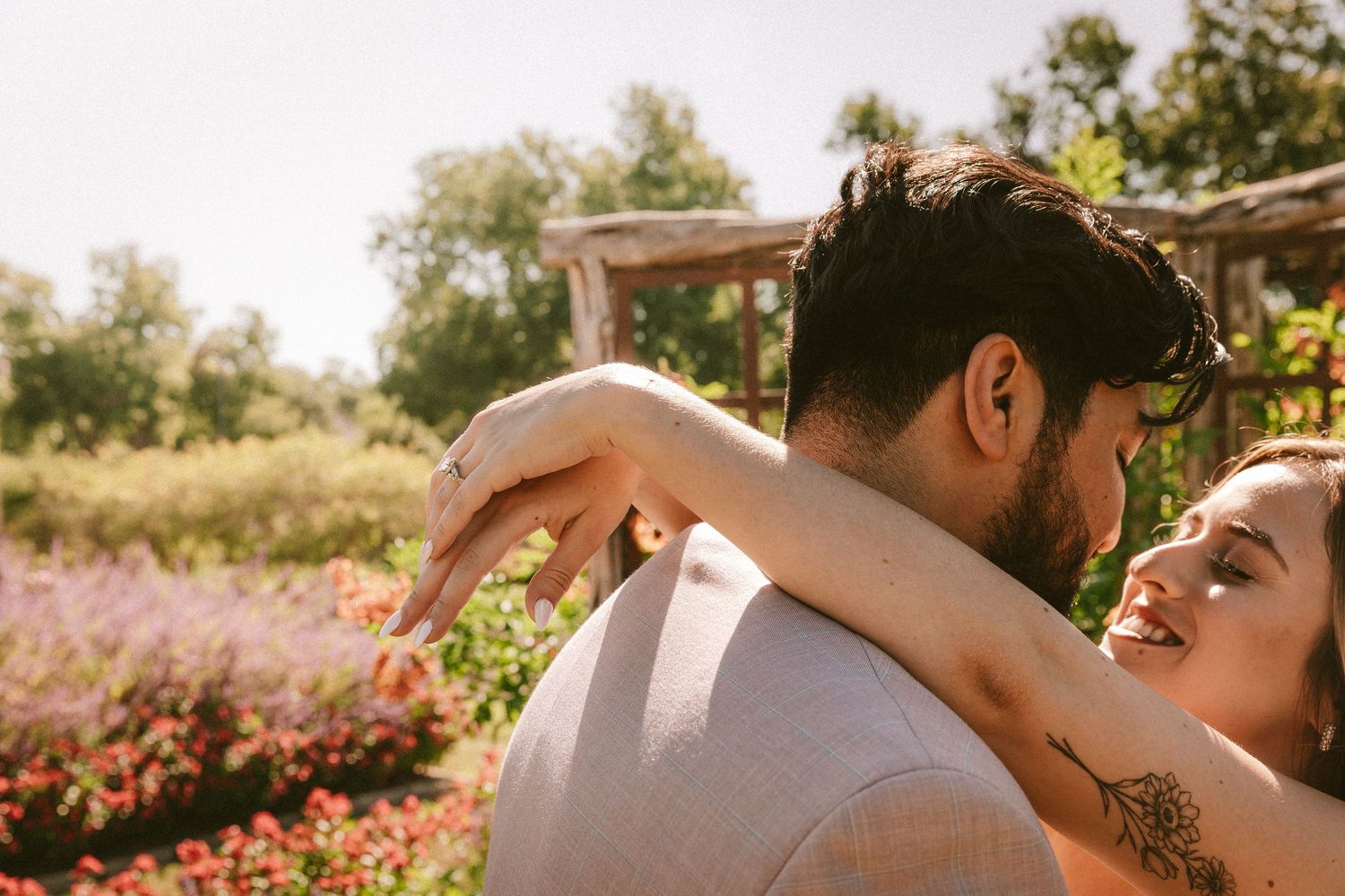 A romantic couple shares an intimate moment in a sunlit garden with pink flowers in the background.