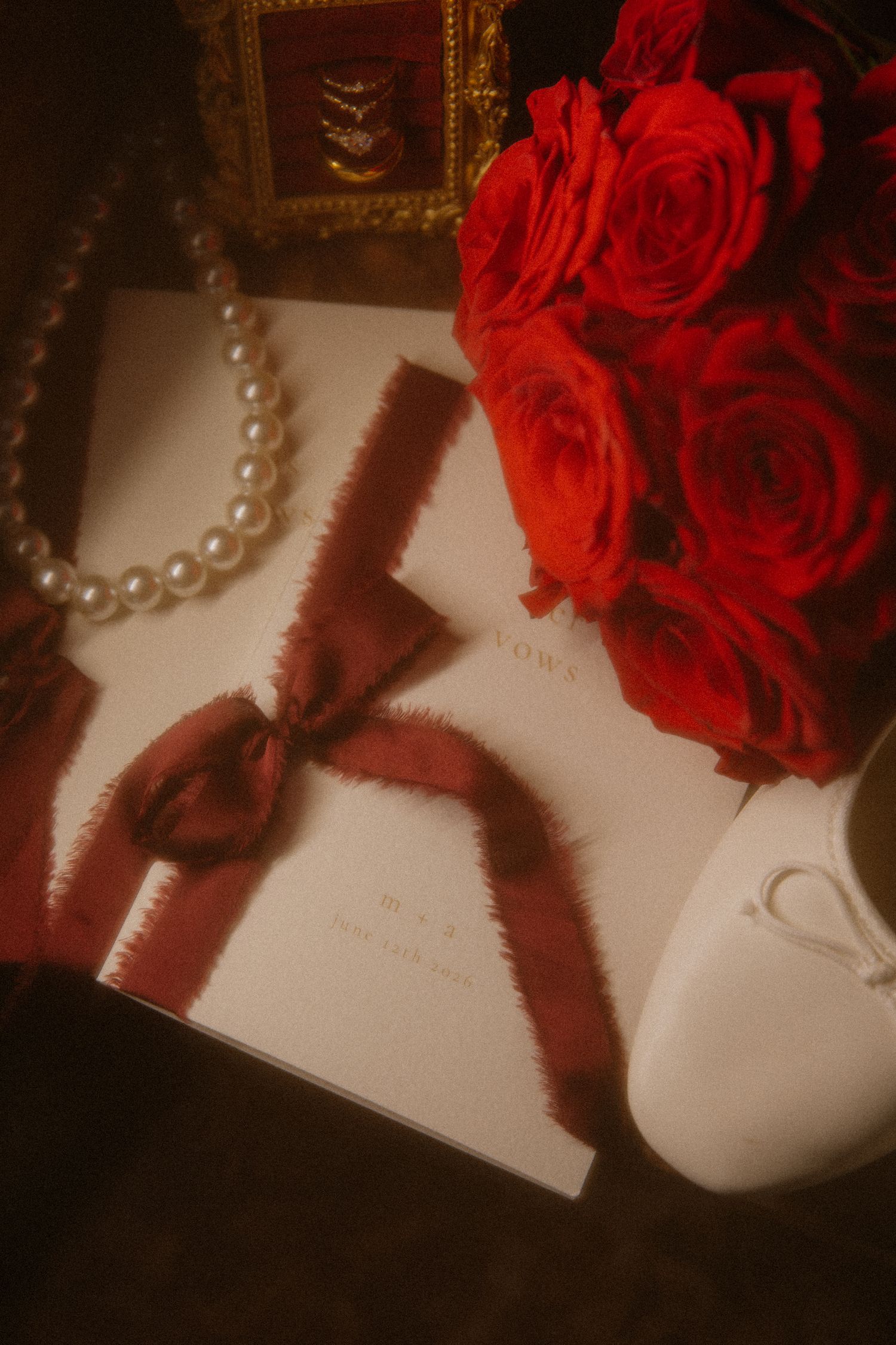 Wedding flat lay with red roses, pearl necklace, burgundy ribbon bow, and white vow book.
