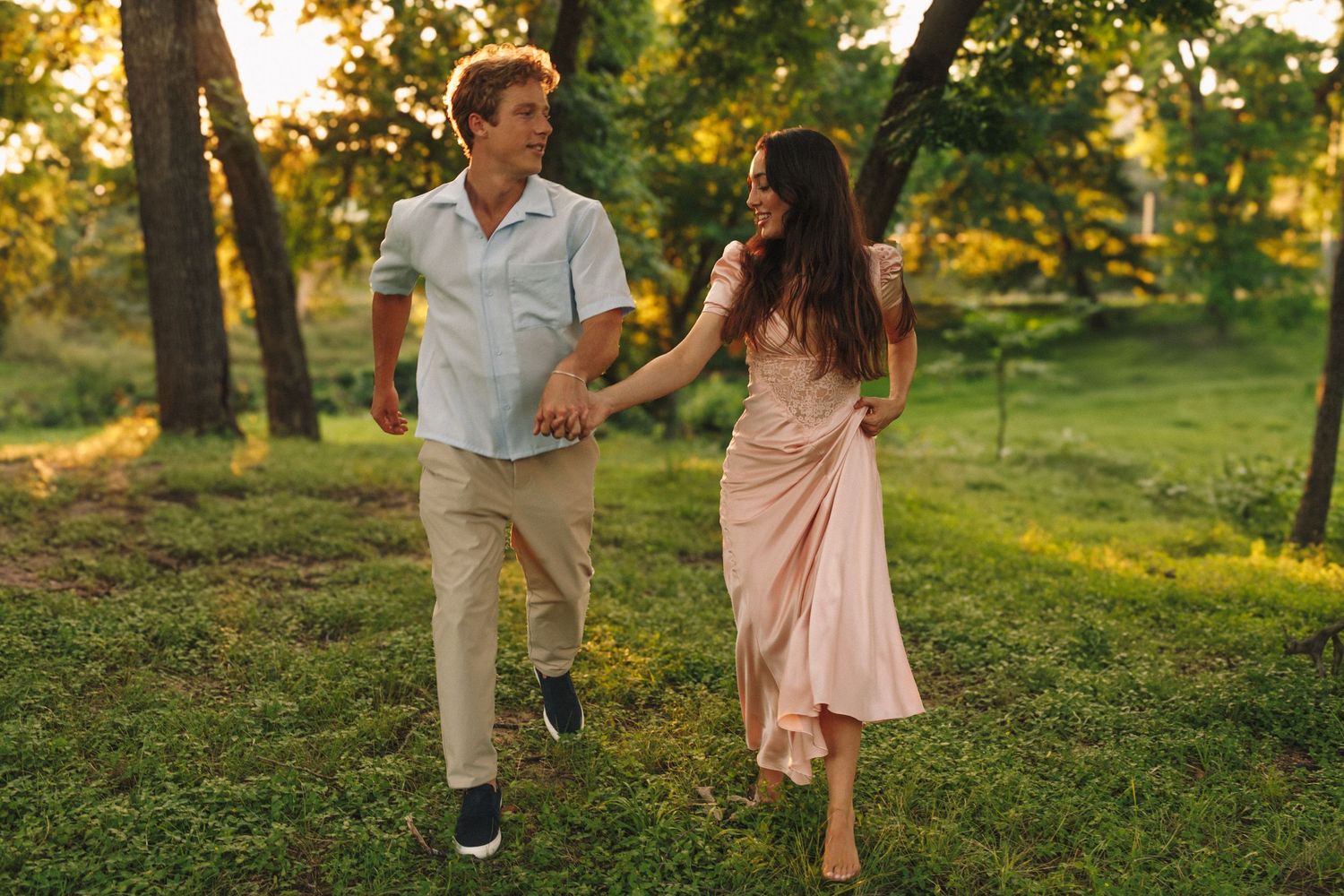 Couple walking together through a sunlit park at golden hour holding hands in romantic summer setting.