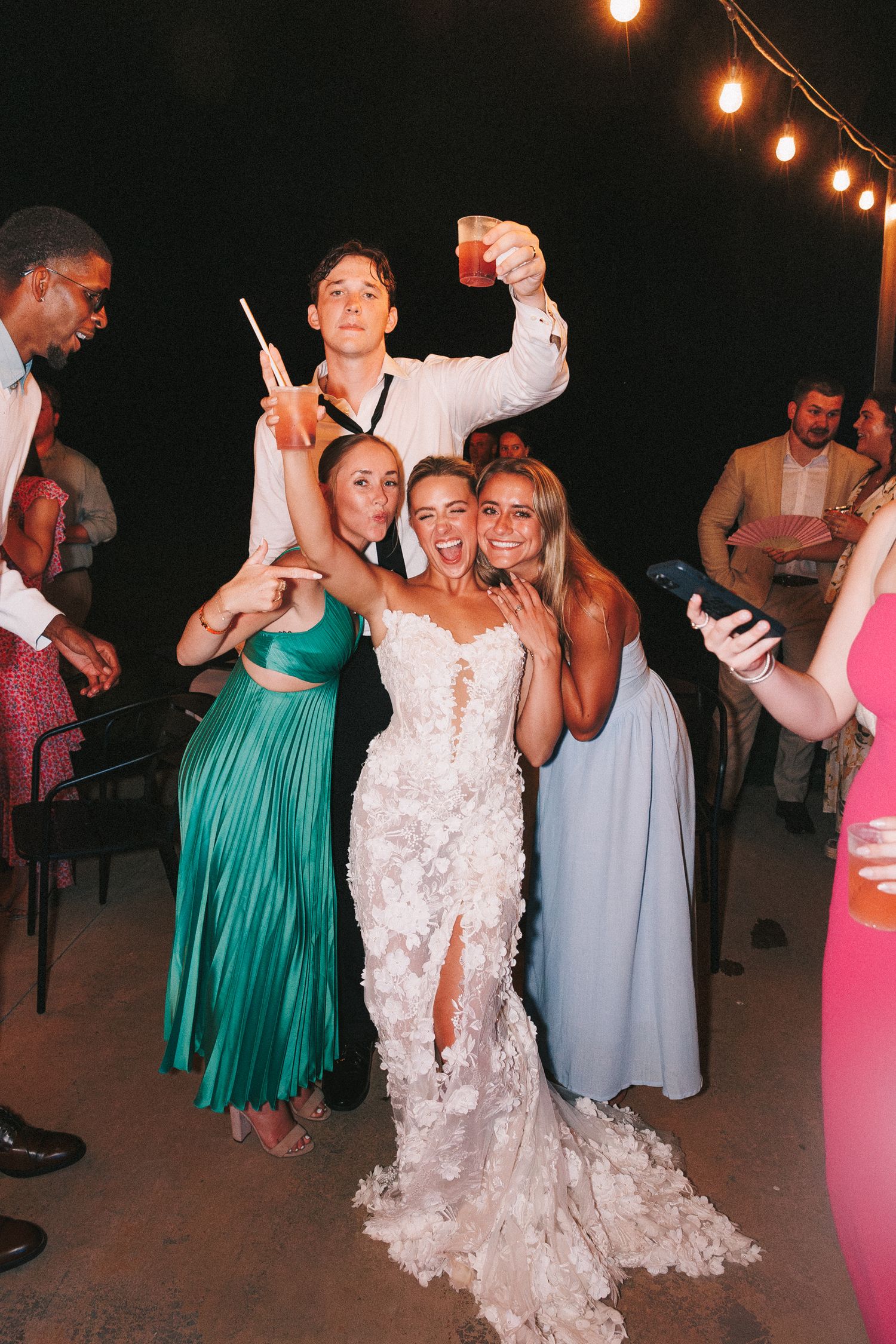 Wedding party celebrates on dance floor under string lights with bride in lace gown and guests in colorful formal attire.
