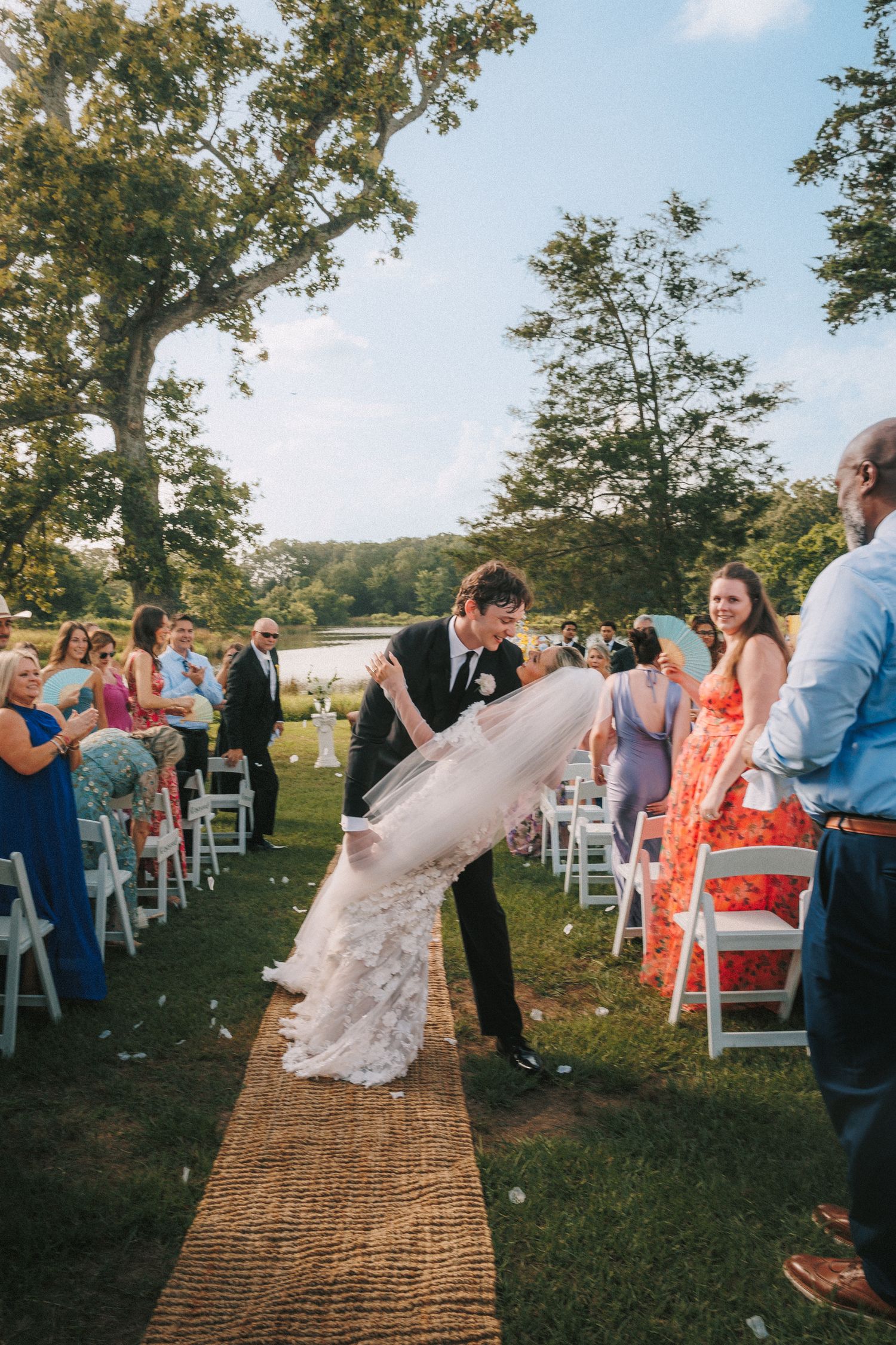 A couple shares a romantic dip kiss at an outdoor wedding ceremony while guests look on from white chairs.