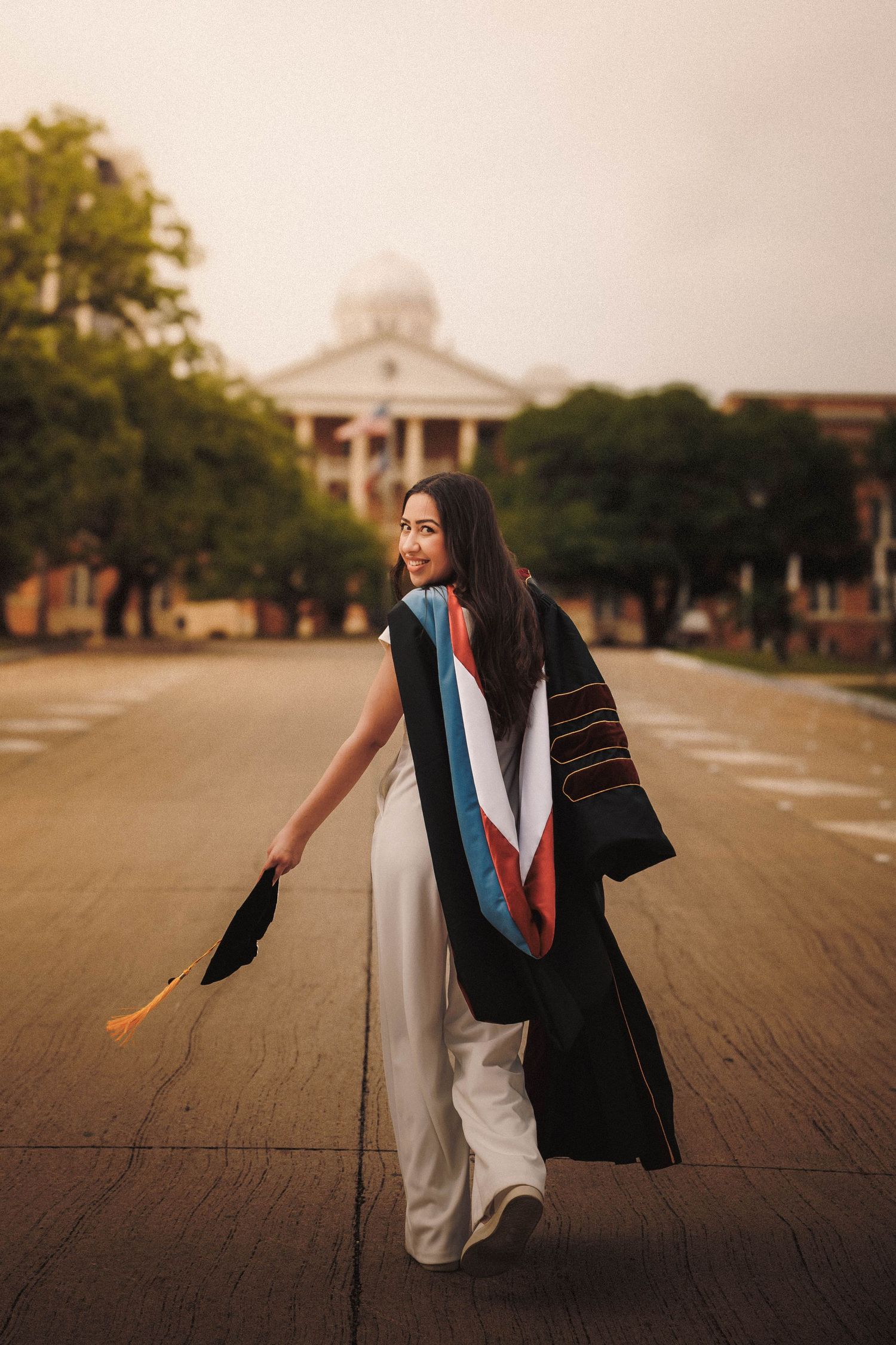 A graduate in a black cap and gown strikes a candid pose on a campus sidewalk during golden hour photography.