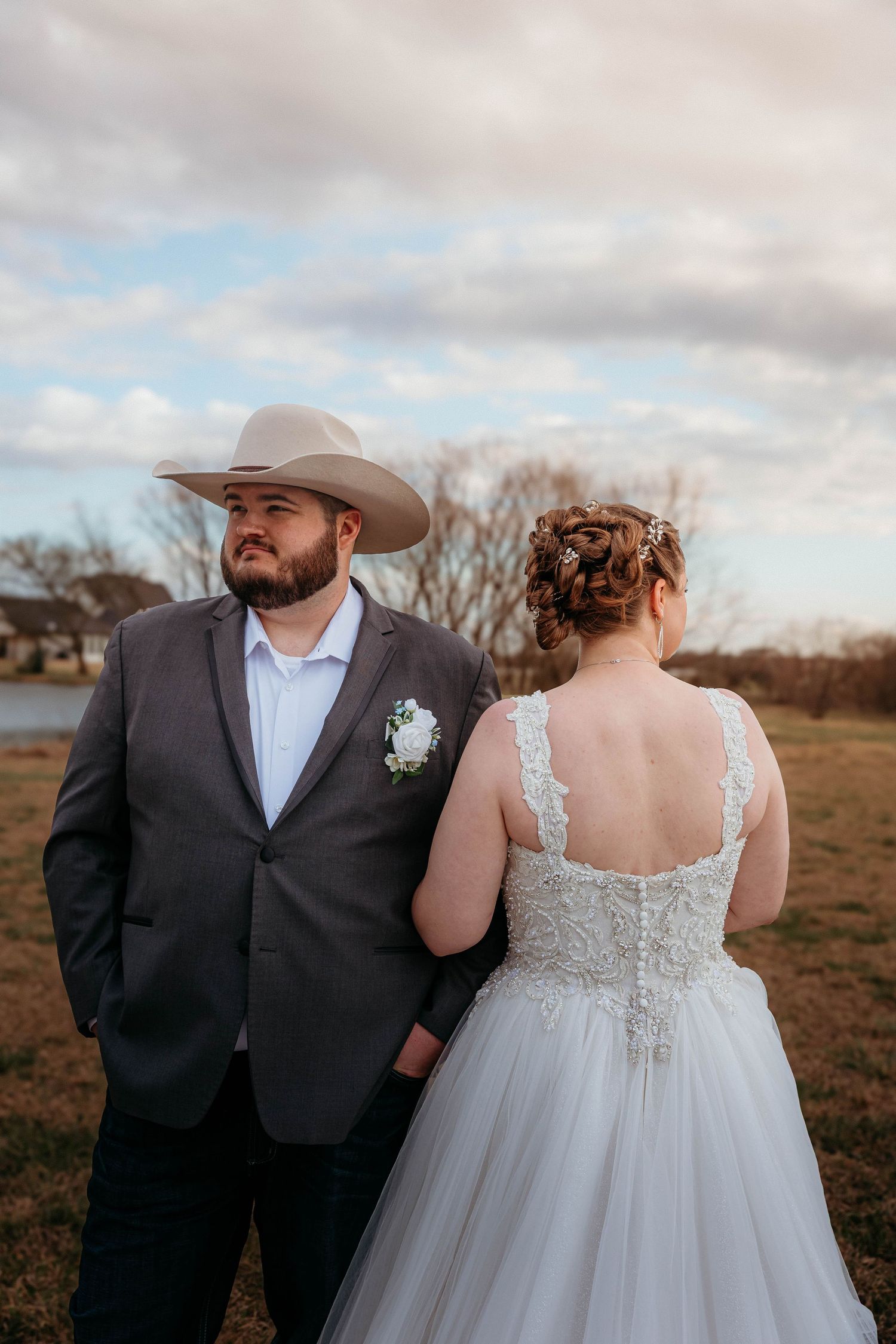 A couple in wedding attire stands in a field with cloudy skies, sharing a romantic moment during their special day.