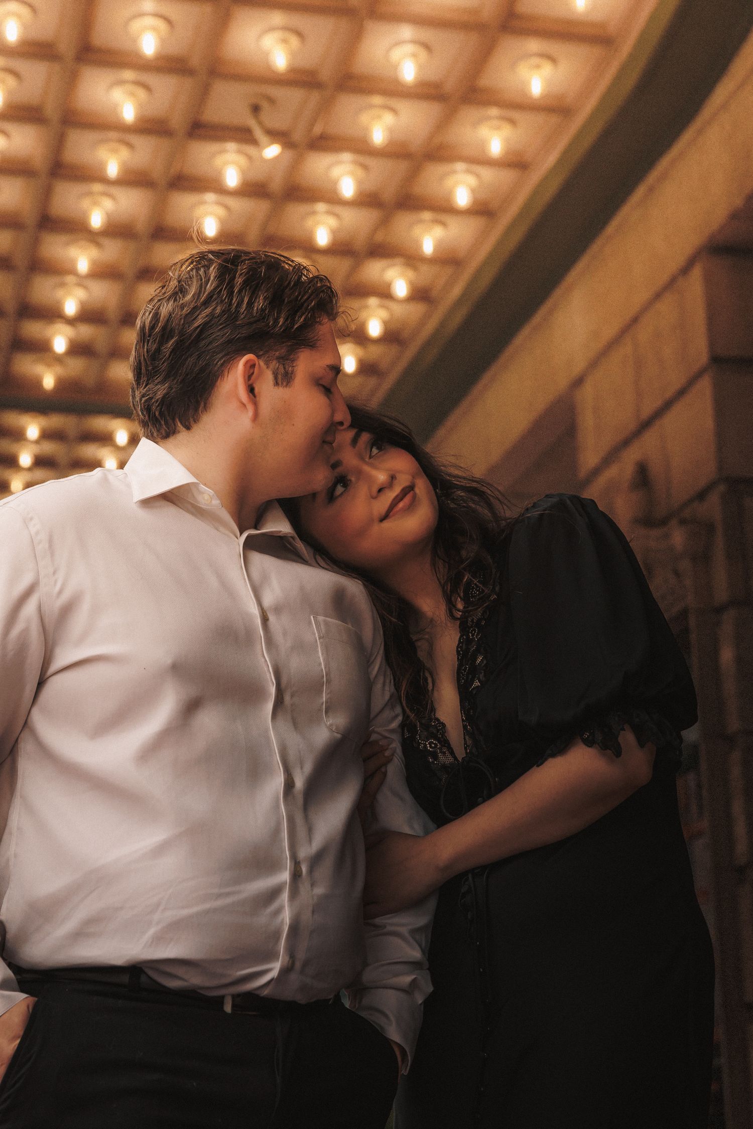 A romantic couple shares an intimate moment under warm marquee lights, man in white shirt kissing woman in black dress.