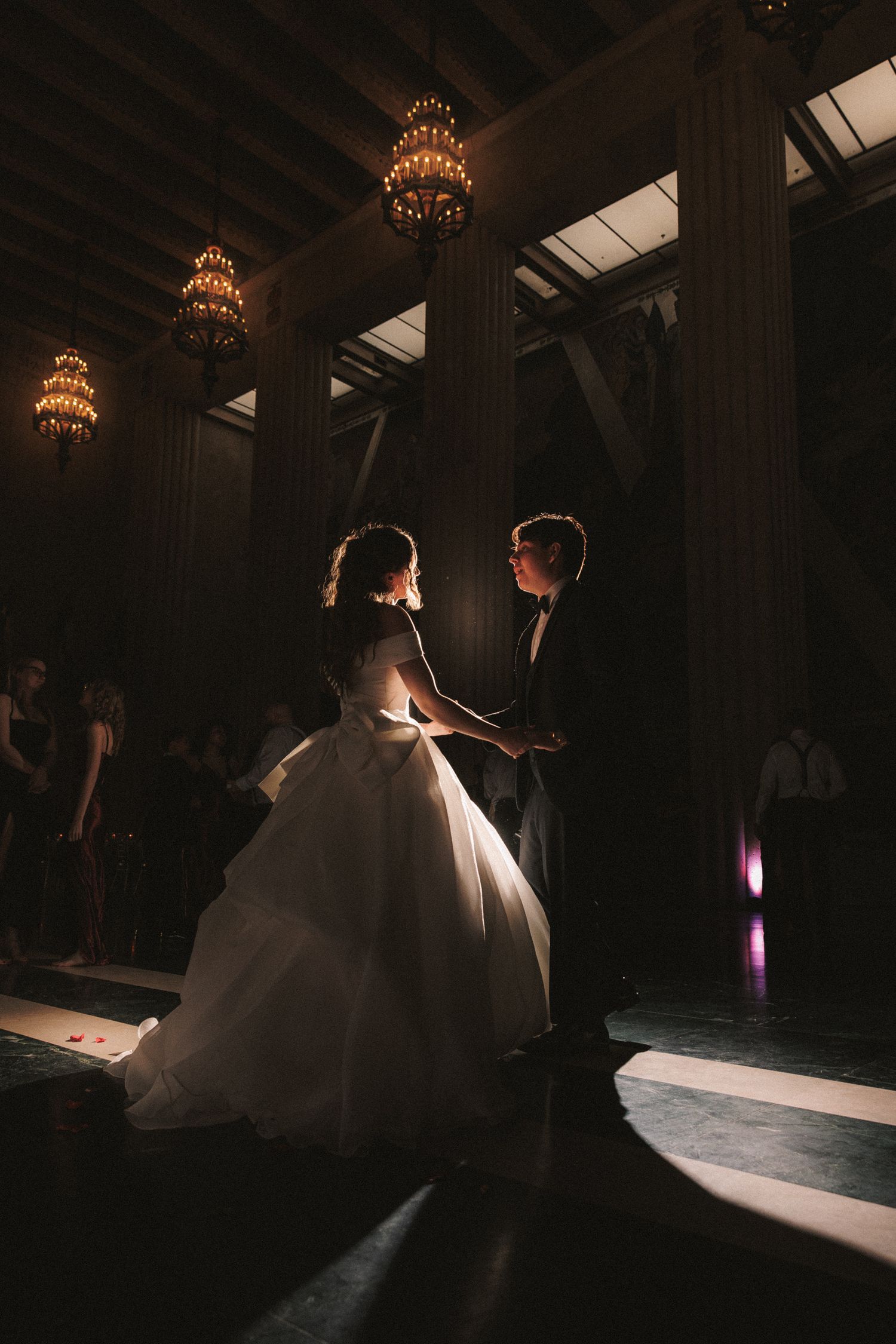 Bride and groom share their first dance in an elegant dimly lit ballroom with chandeliers and wedding guests.