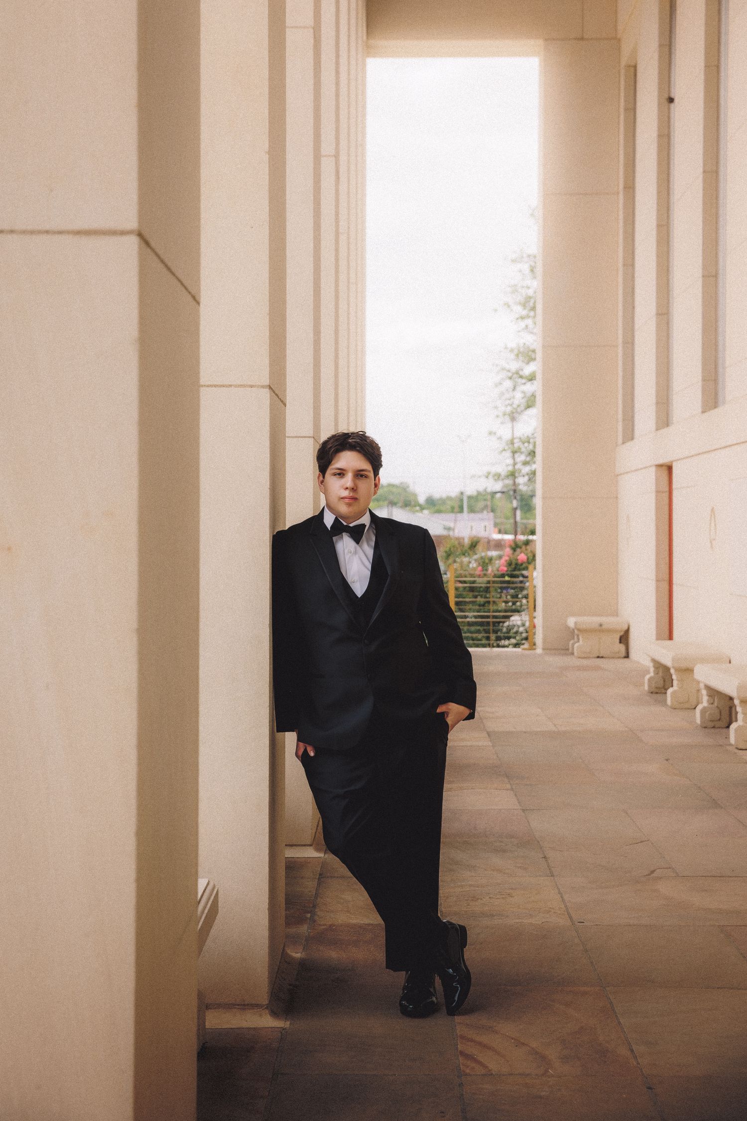 Young man in black tuxedo with bow tie leaning against column in elegant architectural corridor.