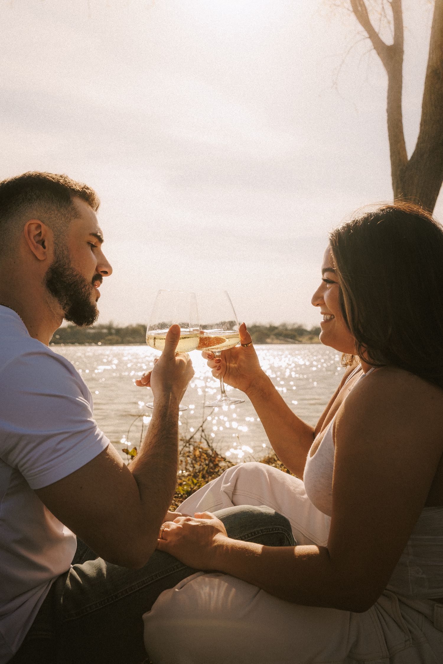 Couple toasting drinks at sunset by the waterfront with sparkling water in background.