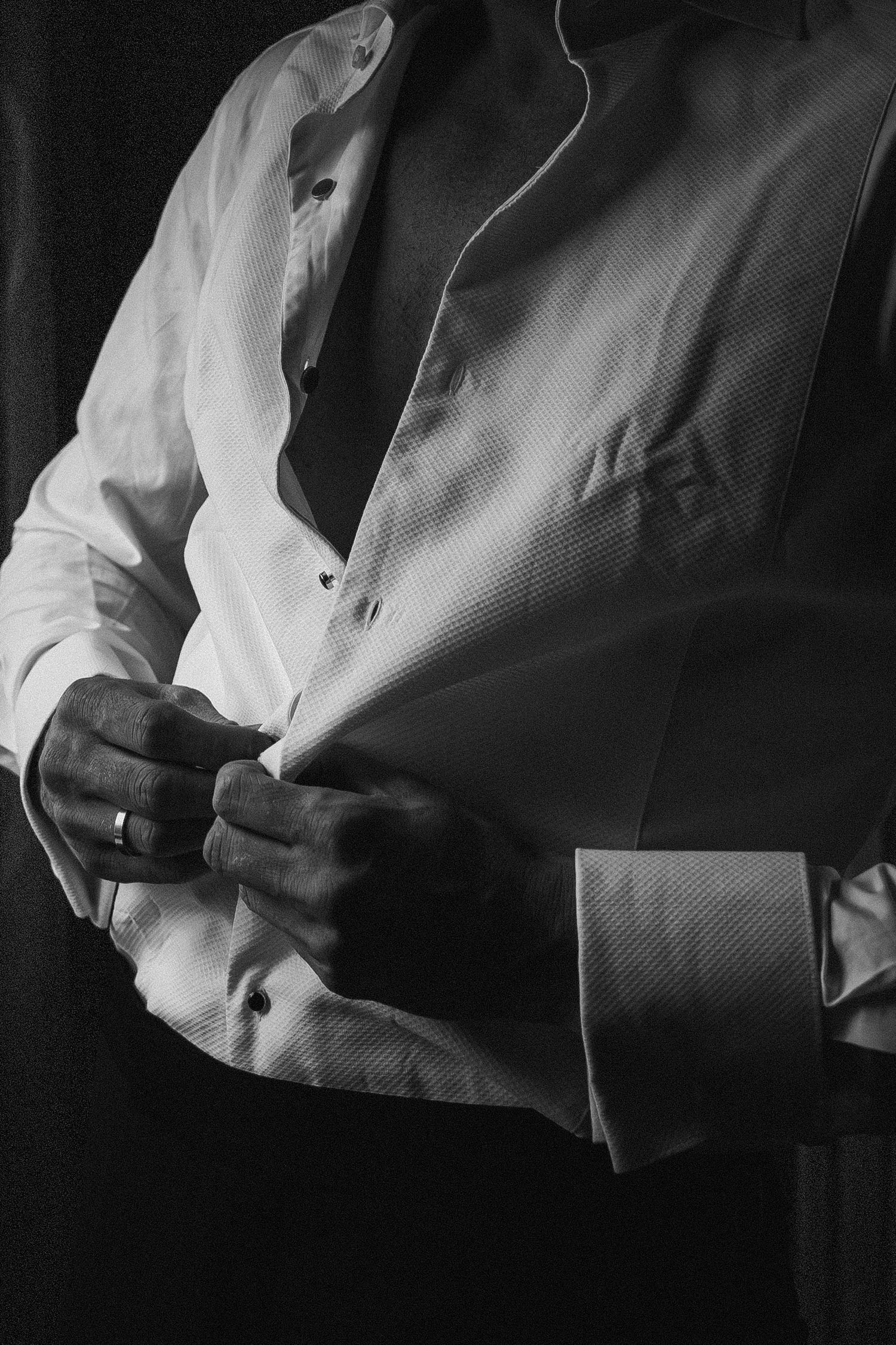 Black and white close-up of groom buttoning white tuxedo shirt before wedding ceremony.