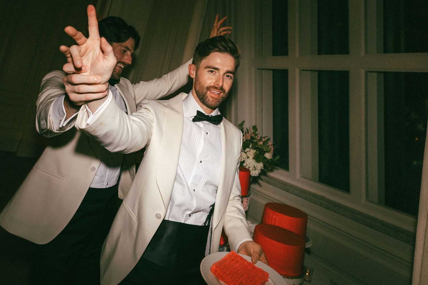 Groom in white tuxedo poses playfully beside red wedding cake at reception.