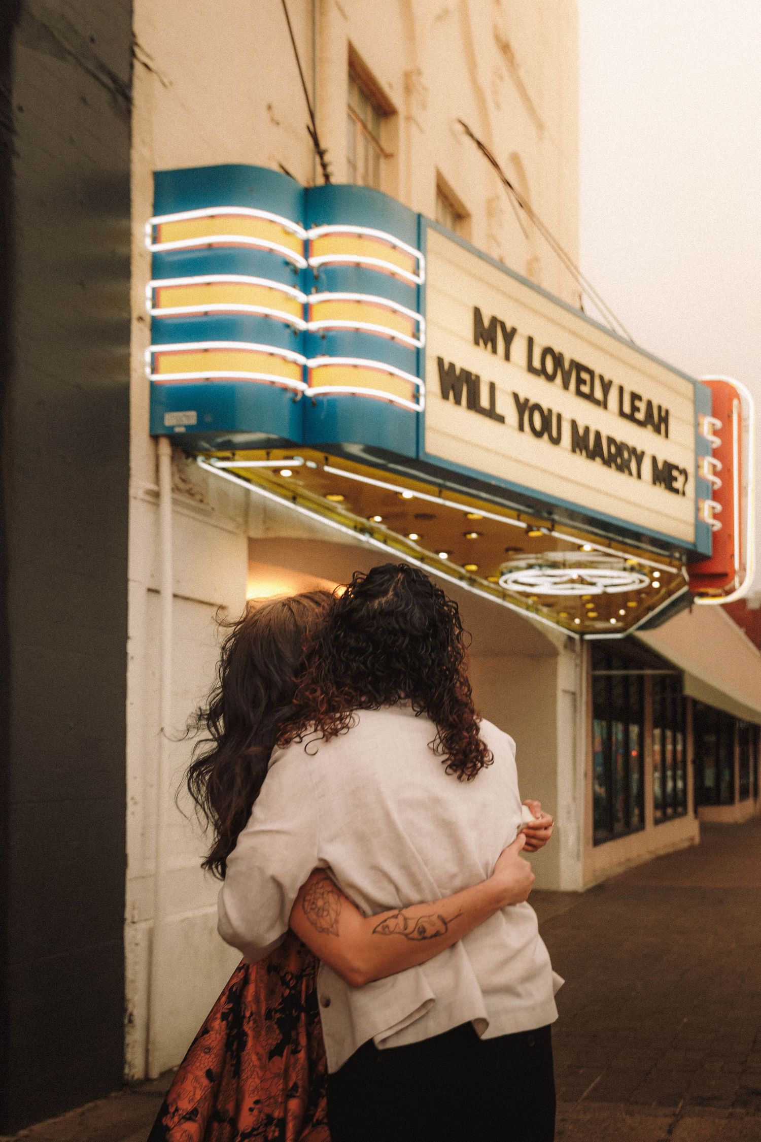 Couple embracing under a theater marquee displaying a romantic marriage proposal message.