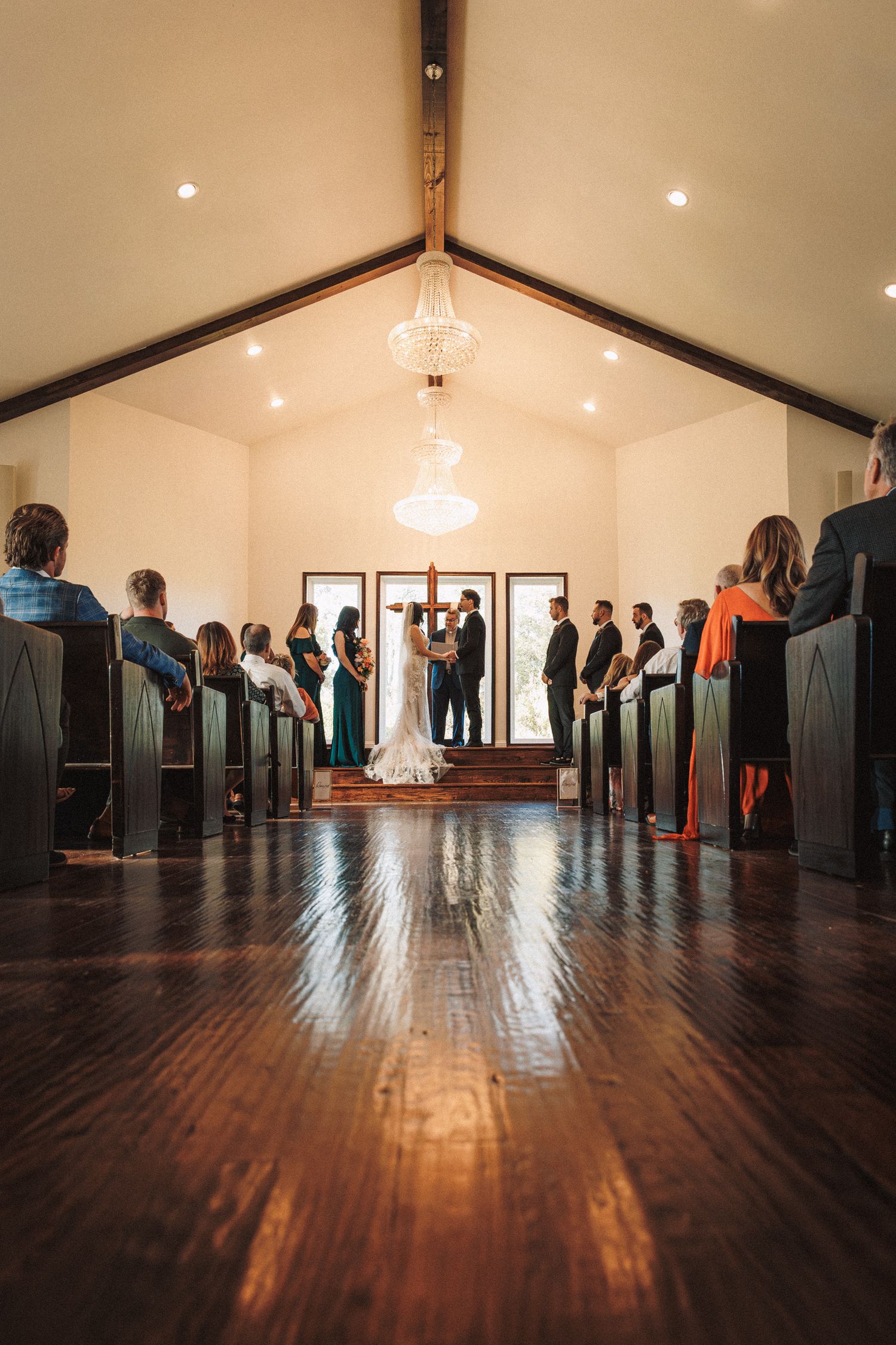 Wide-angle view of a wedding ceremony in a chapel with dark wood pews and a chandelier overhead.
