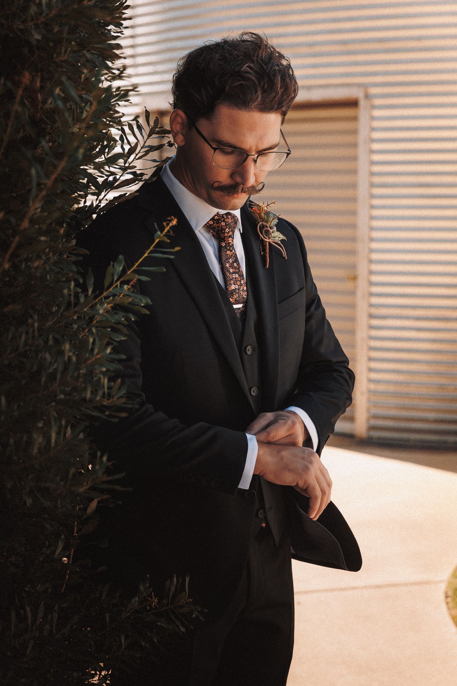 Groom in dark suit adjusting cufflinks outdoors with floral boutonniere and patterned tie in warm sunlight.
