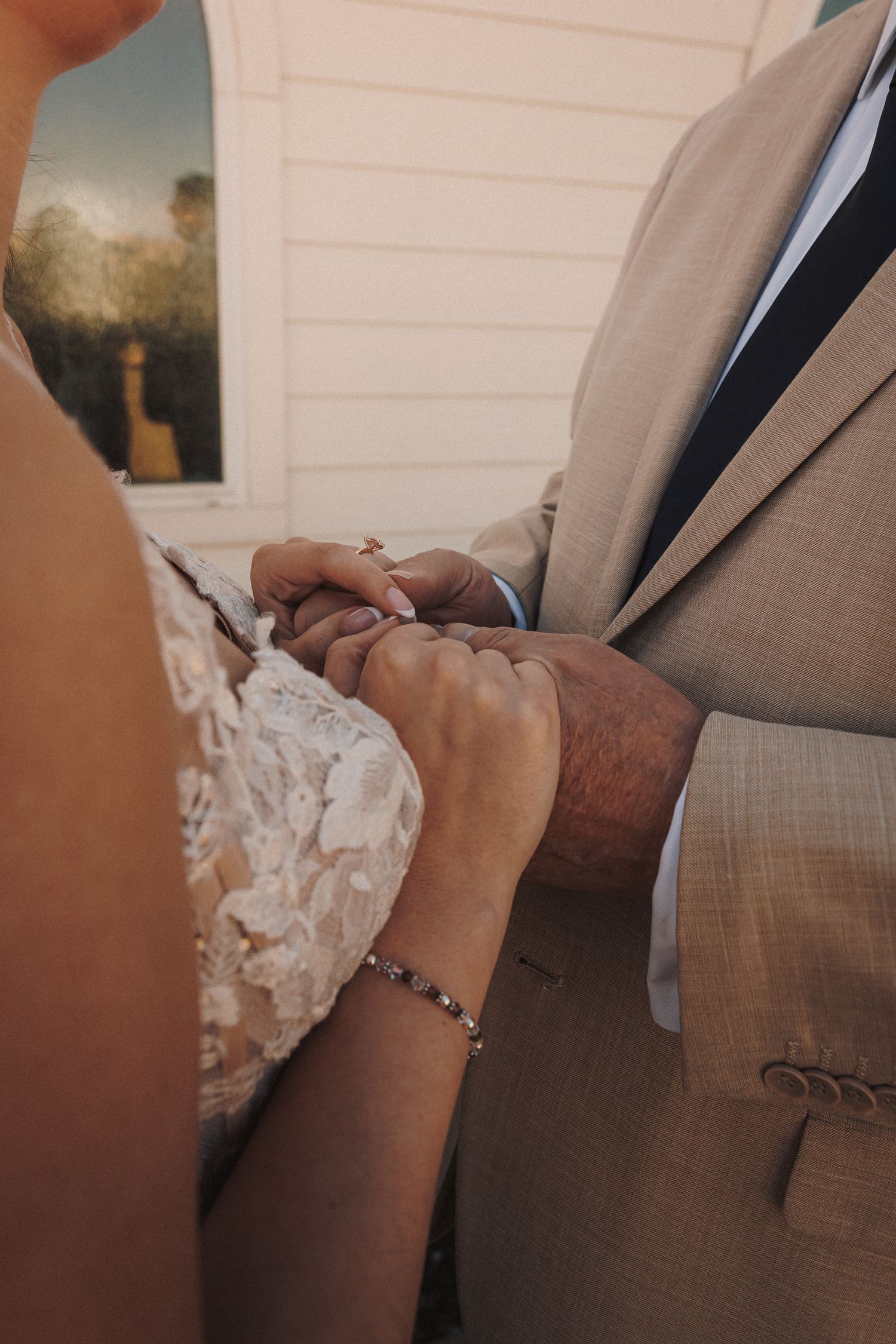 Bride and groom exchange rings during outdoor wedding ceremony, close-up of hands with lace dress and bracelet.