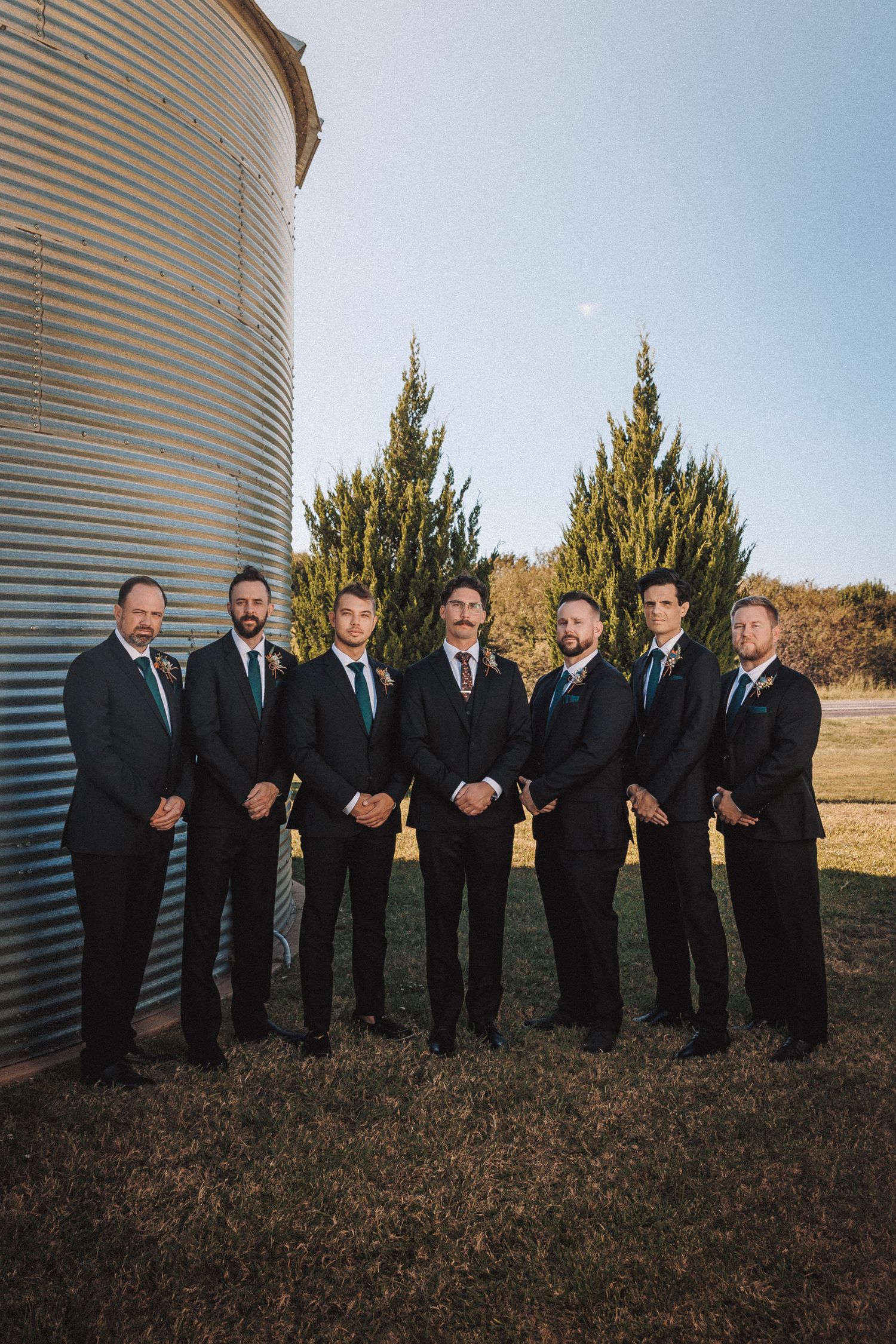 Groom and groomsmen in black suits posing beside a grain silo outdoors.