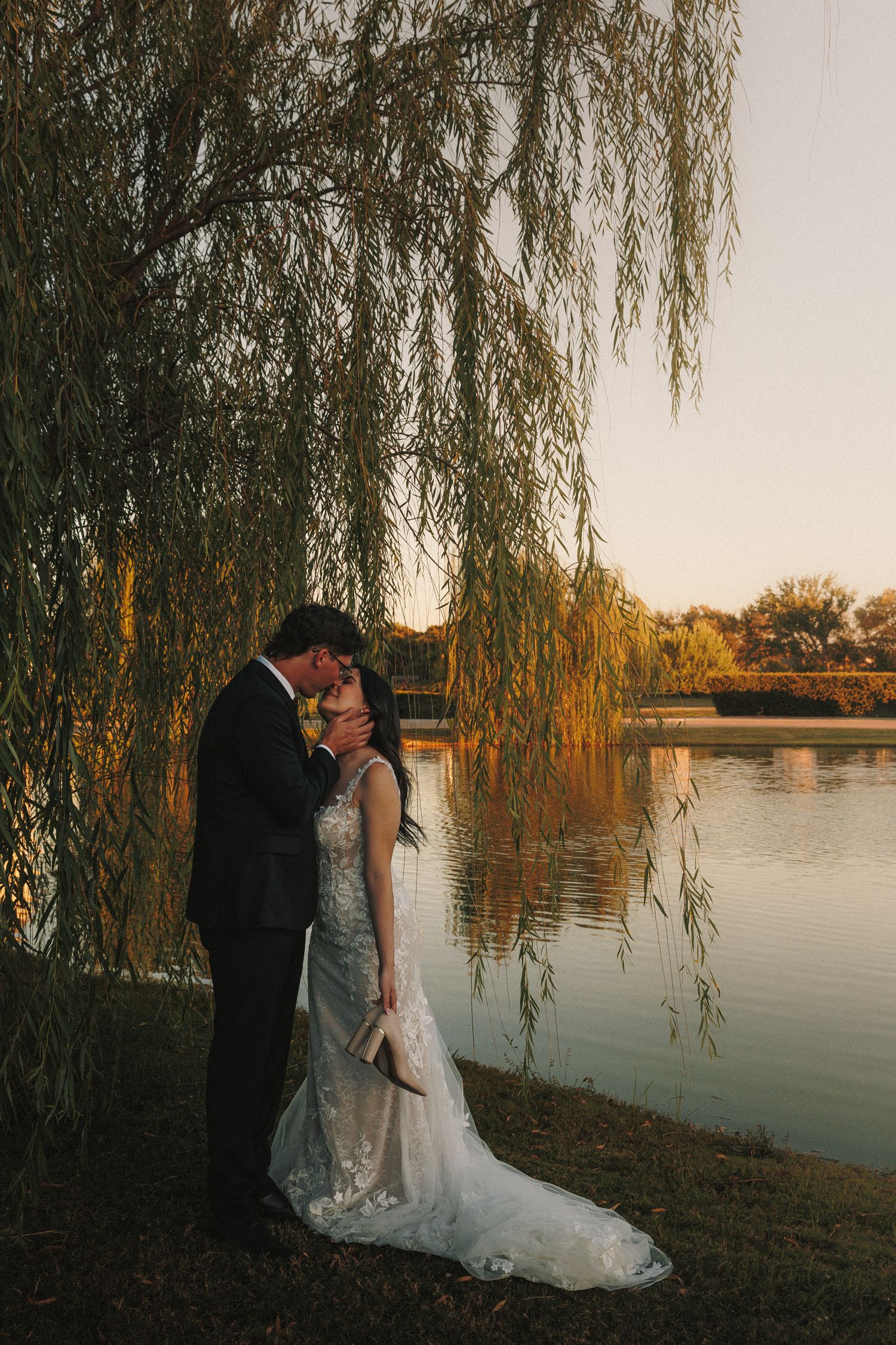 Bride and groom embracing under weeping willow tree by lake at golden hour sunset.