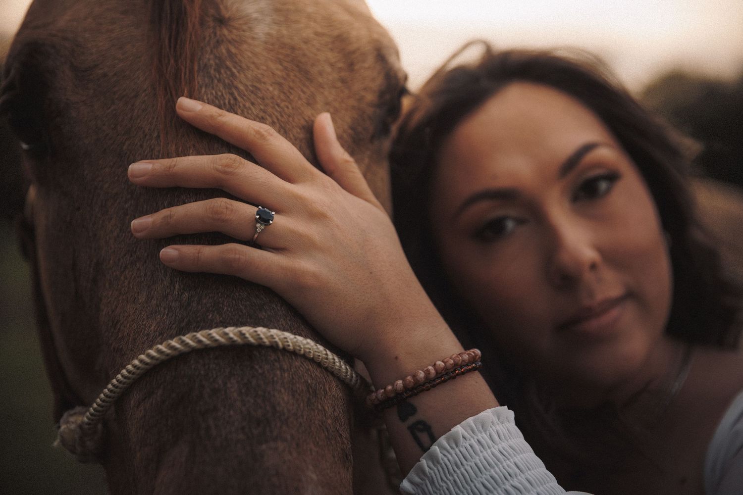 Close-up of a woman with an engagement ring resting her face against a brown horse at sunset.